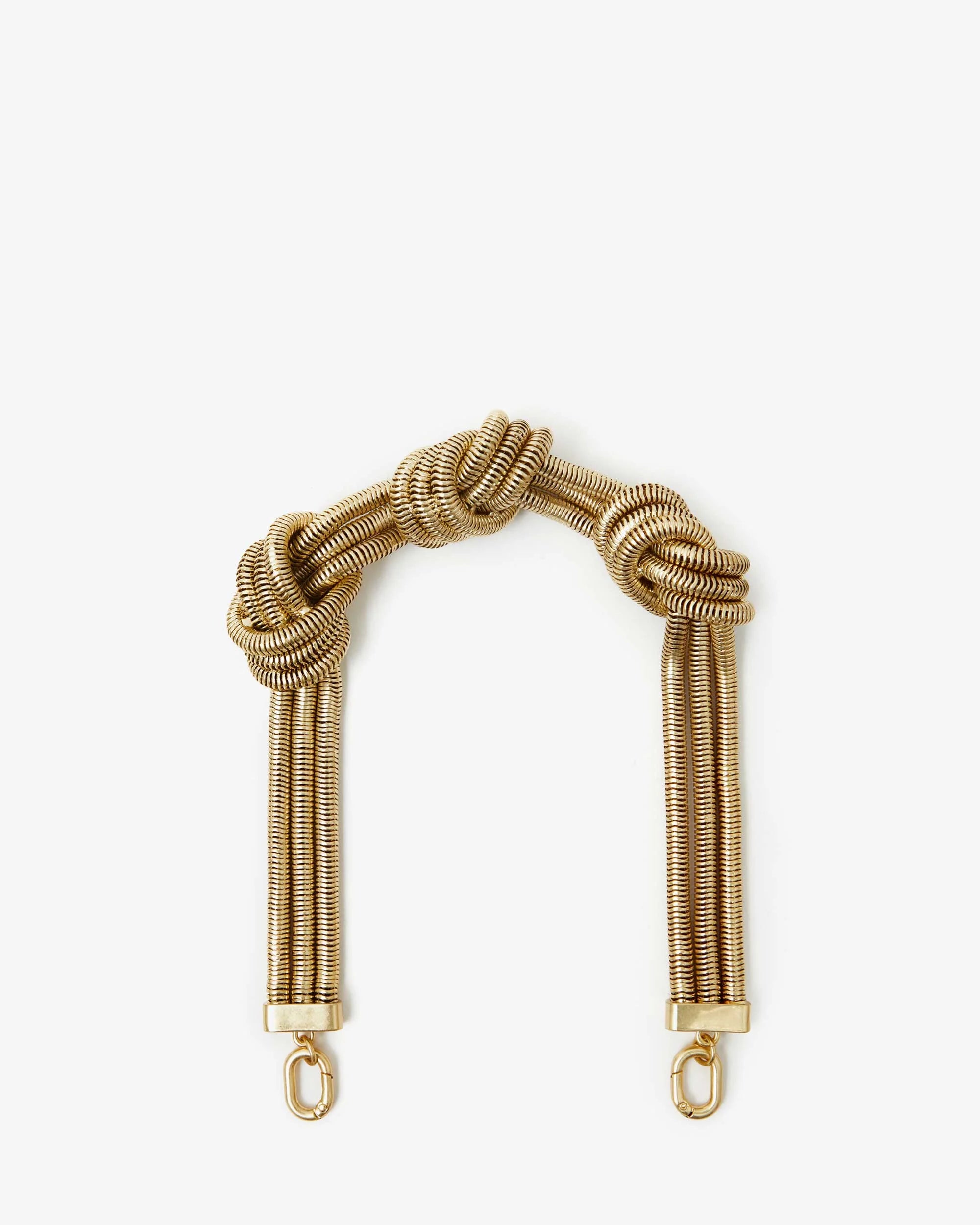 The Shortie Strap by Clare Vivier is an elegant gold-tone metal handbag strap with a coiled design and decorative knots, featuring lobster claw clasps for easy attachment. It is showcased against a white backdrop.