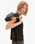 A woman with shoulder-length blonde hair wears a black t-shirt and jeans, holding a black woven handbag featuring the Clare Vivier Shortie Strap/Mariner Chain over her shoulder, looking at the camera against a white background.
