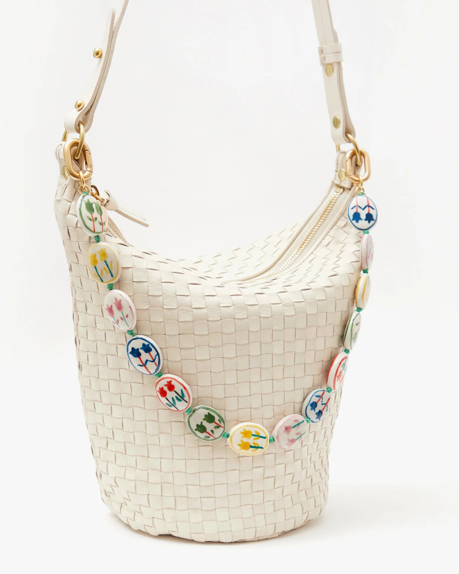 The Petit Jeanne by Clare Vivier is a white handwoven leather bucket bag with gold zipper and strap hardware, a removable shoulder strap, and a colorful circular charm strap adorned with floral designs.