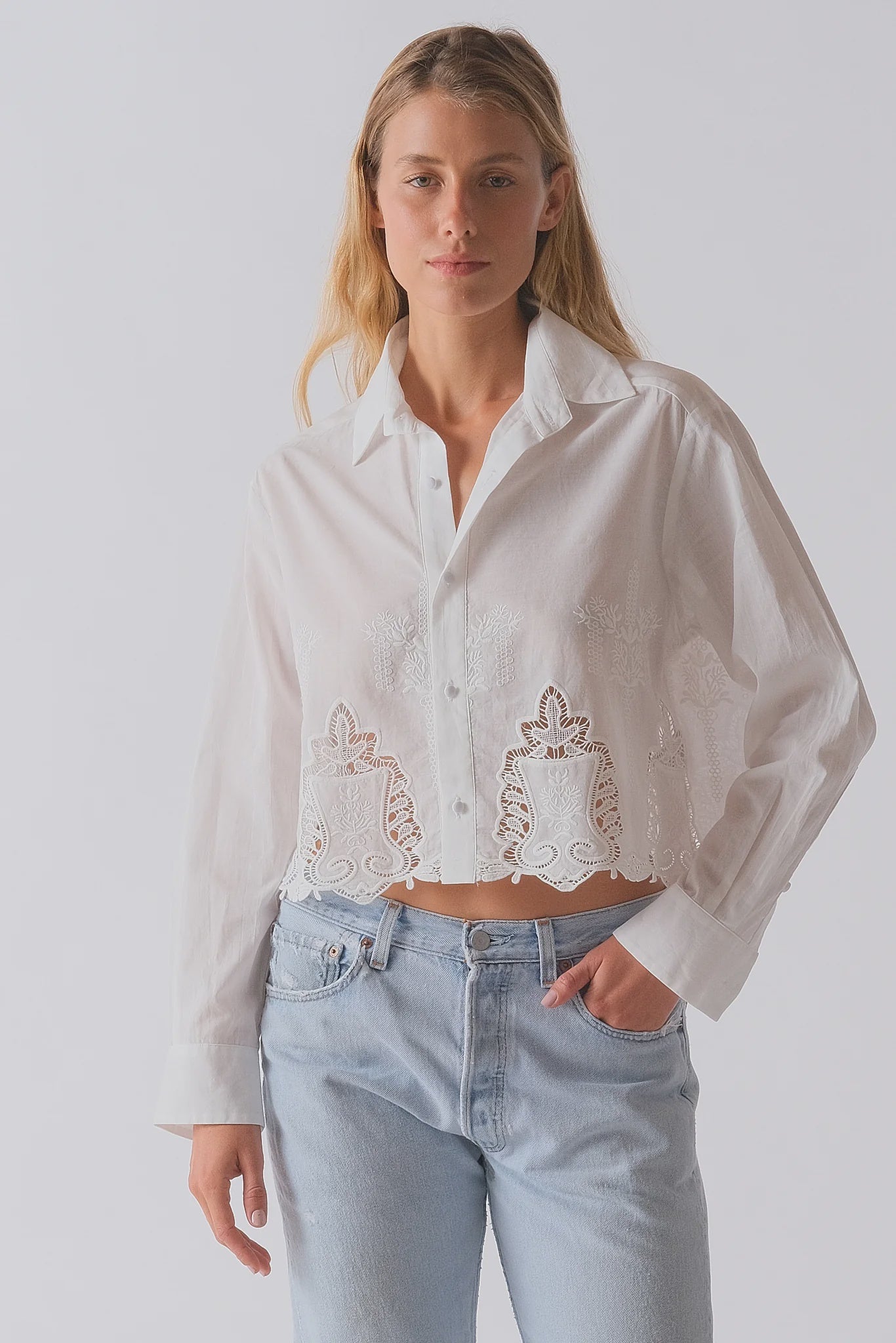 A woman with light hair wears Cissa's The Shorty Shirt—a white, 100% cotton, long-sleeved collared blouse with embroidery and sheer bottom details—paired with light blue high-waisted jeans against a plain background.