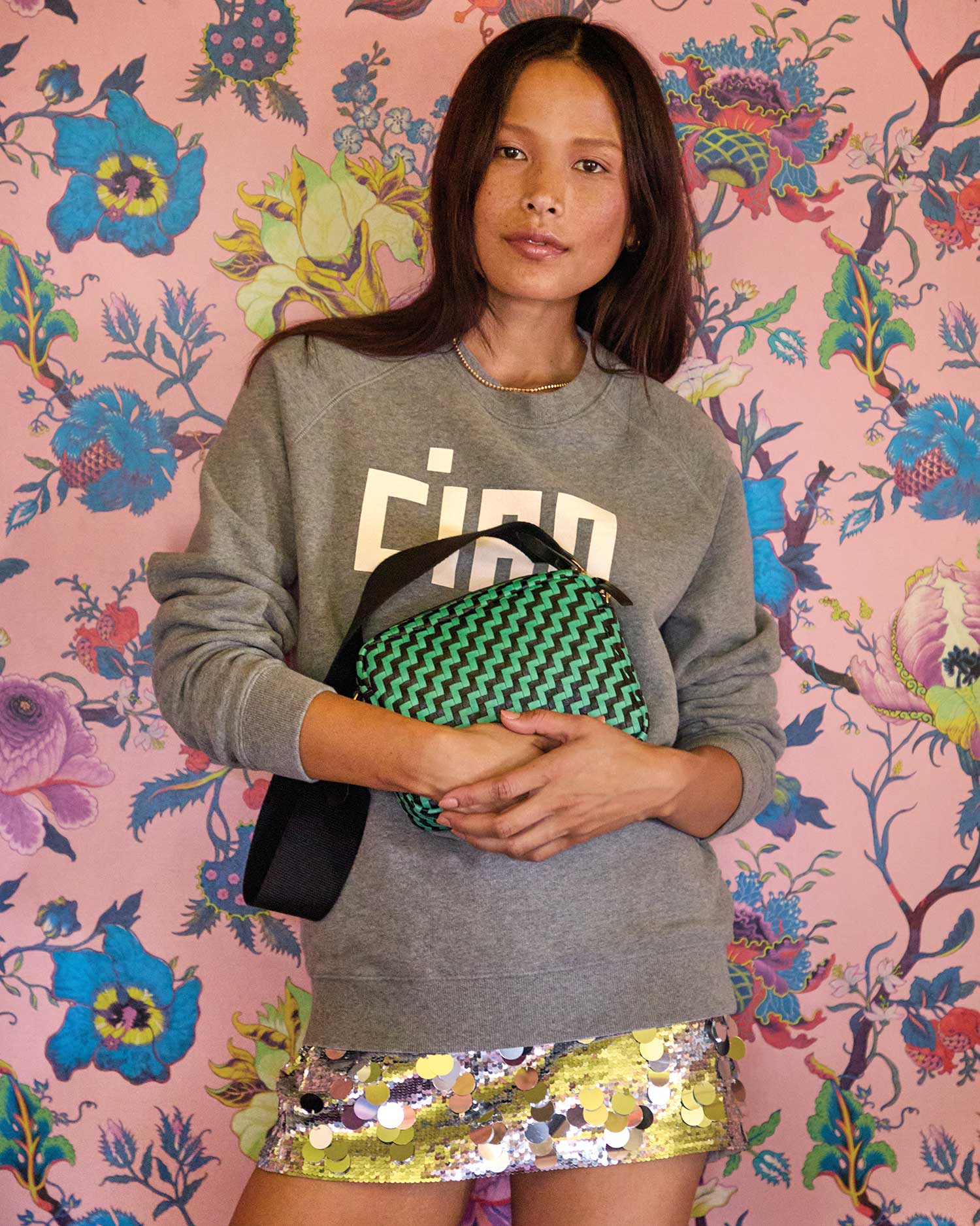 A person stands against a vibrant floral background, wearing a gray sweater with bold text and a sequined skirt. They carry the Clare Vivier Shoulder Strap, featuring a green and black pattern with a cotton webbing strap, exuding a casual yet stylish look that's undeniably Made in L.A.