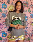 A person stands against a vibrant floral background, wearing a gray sweater with bold text and a sequined skirt. They carry the Clare Vivier Shoulder Strap, featuring a green and black pattern with a cotton webbing strap, exuding a casual yet stylish look that's undeniably Made in L.A.