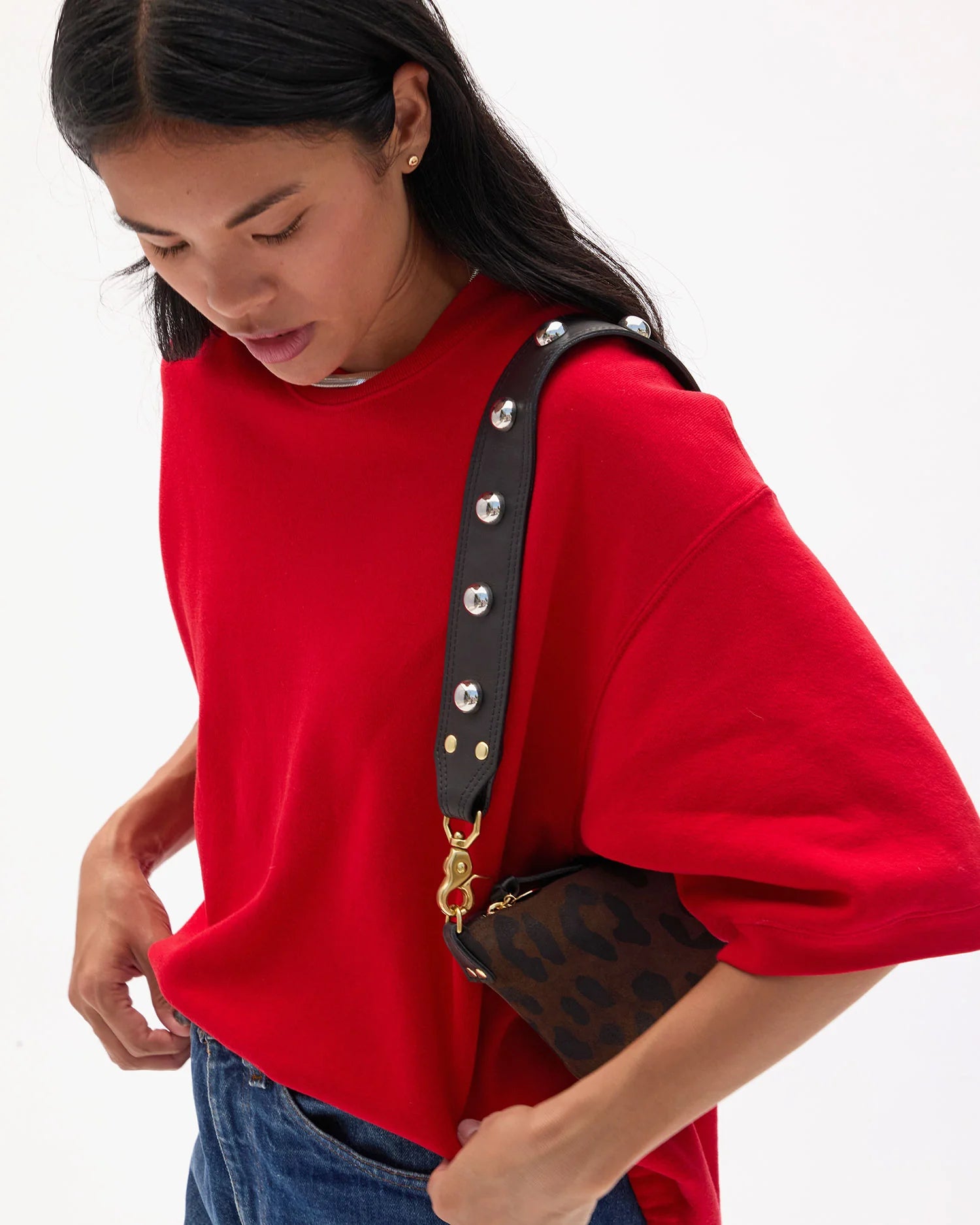 A woman in a loose red top and blue jeans looks down, carrying the Clare Vivier Shoulder Strap Naturelle W/ Studs—a small brown leopard-print bag with a black leather strap accented by shiny brass studs—against a plain white background.