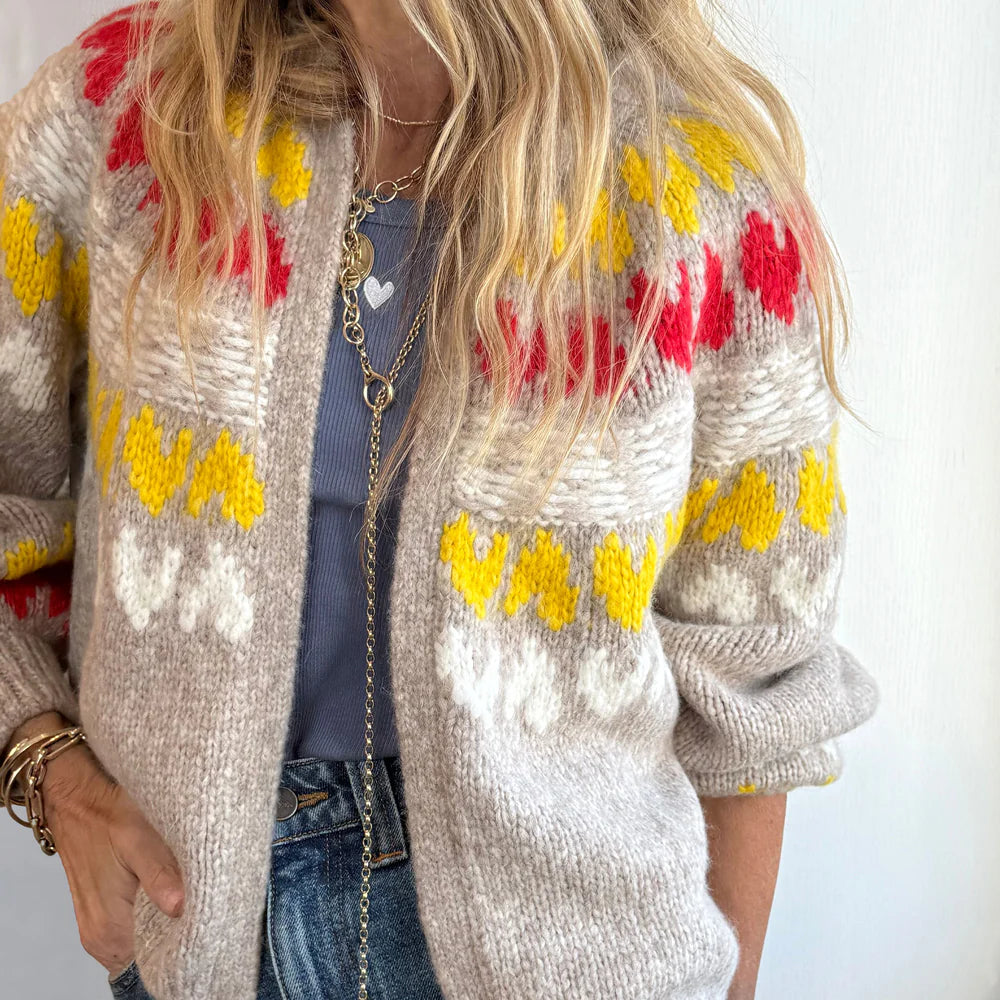 A woman styles the Kerri Rosenthal Sienna Ker-isle Cardigan, a chunky knit featuring yellow, red, and white Fair Isle-inspired patterns, with a blue top, jeans, wavy blonde hair, layered necklaces, and bracelets.