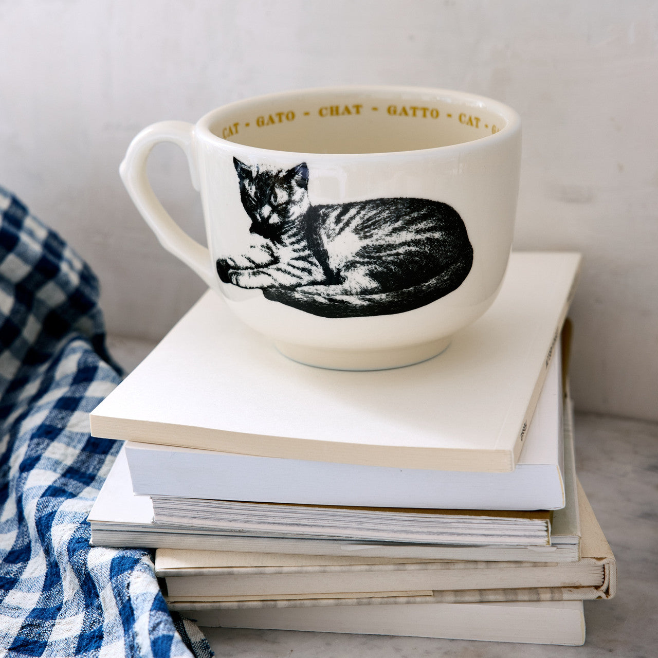 The Sir/Madam Grand Cup, white with a black cat illustration and "CAT," "GATTO," and "CHAT" around the rim, rests atop books beside a blue and white checkered cloth.