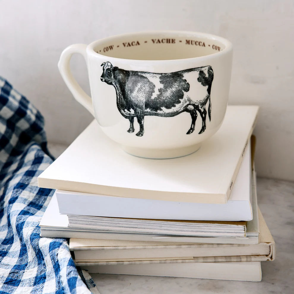 The Grand Cup by Sir/Madam, a large white cup featuring a black cow illustration and &quot;cow&quot; in multiple languages on the rim, sits atop books beside a blue-and-white checkered cloth.