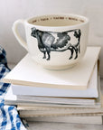 The Grand Cup by Sir/Madam, a large white cup featuring a black cow illustration and "cow" in multiple languages on the rim, sits atop books beside a blue-and-white checkered cloth.