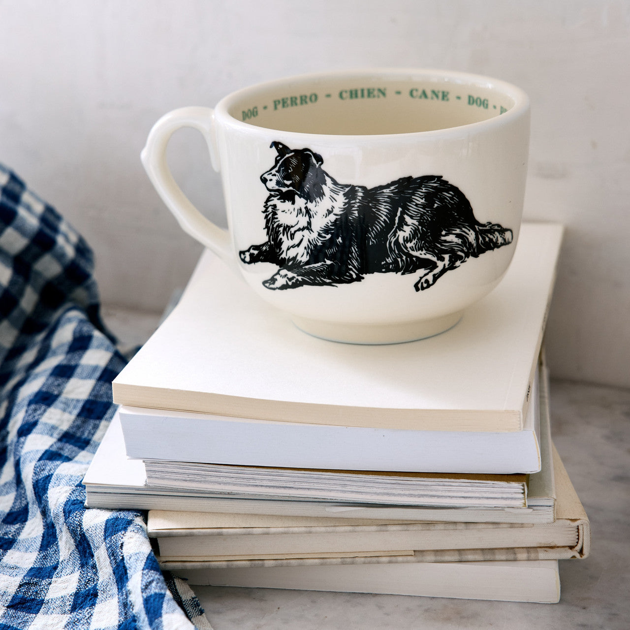 The Sir/Madam Grand Cup, a white mug with a black and white dog illustration, rests on books beside a blue and white checkered cloth. Its rim displays the dog's name in colorful repetition across four languages.
