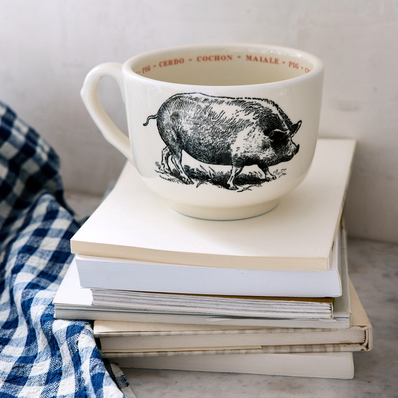 The Sir/Madam Grand Cup, a large white mug with a black pig illustration, rests on stacked books beside a blue and white checkered cloth. The rim features the animal's name in four languages for colorful repetition.