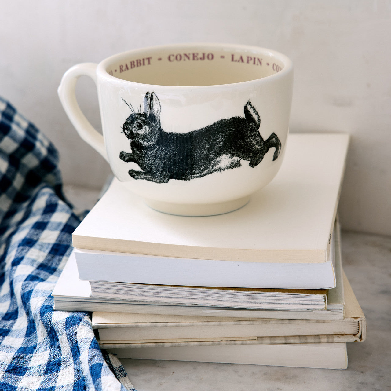 The Grand Cup by Sir/Madam, featuring a black rabbit illustration and &quot;rabbit&quot; in several languages around the rim, sits atop white books next to a blue and white checkered cloth.
