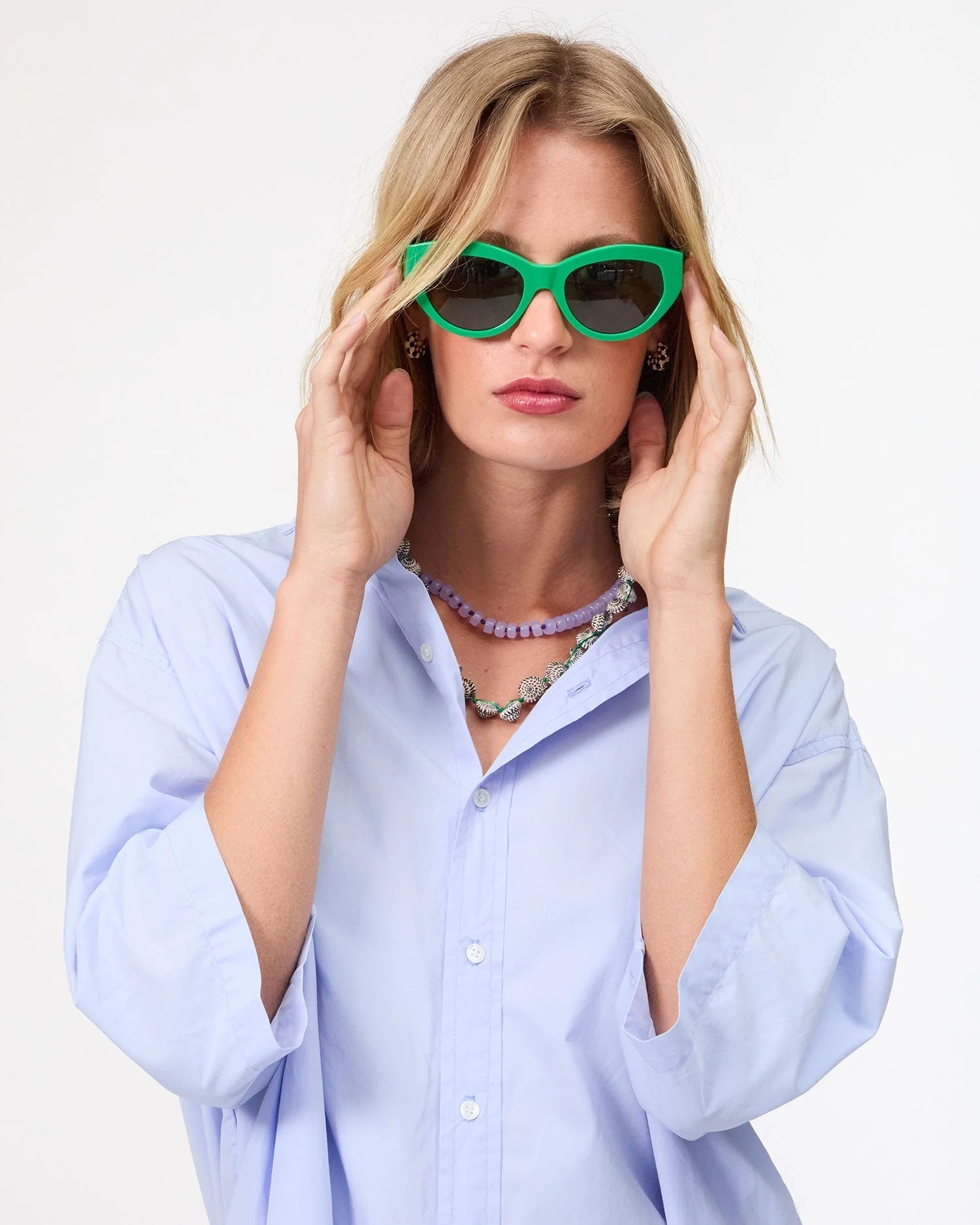 A woman wears bright green cat-eye sunglasses, a light blue button-up shirt, and the Clare Vivier Stone Slice Necklace as she poses adjusting her sunglasses against a plain white background.
