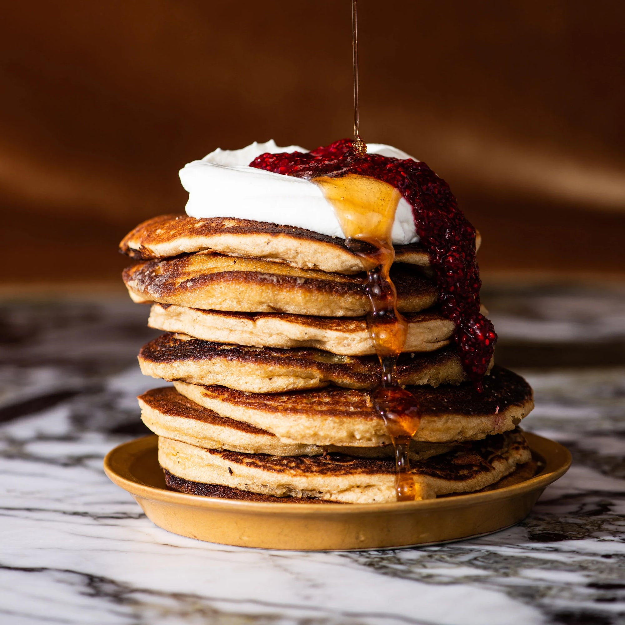 A stack of Wild Rye Almond Flour Pancake Mix pancakes on a golden plate, topped with whipped cream and red berry sauce, with syrup being poured over them, sits on marble against a warm brown background.
