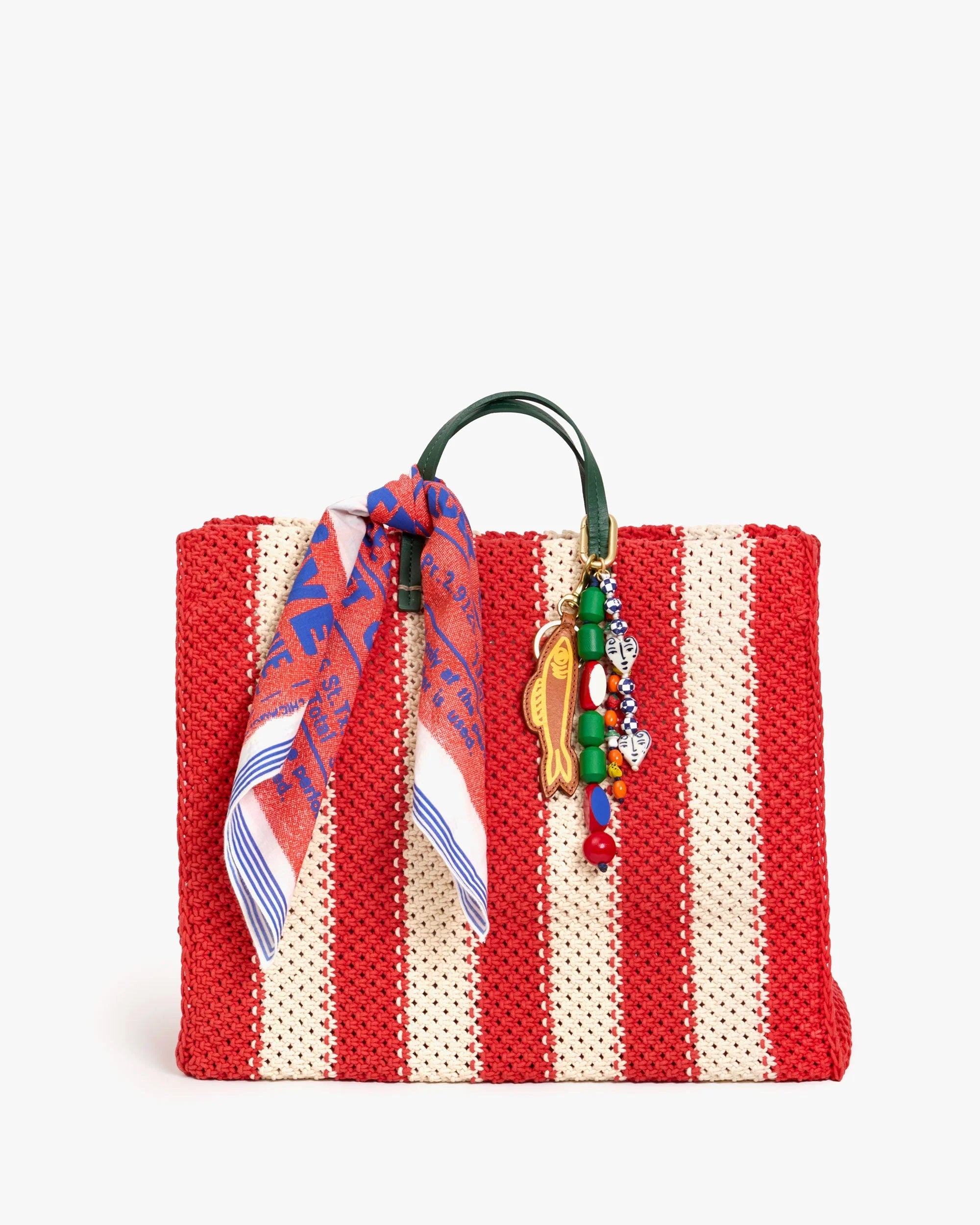 The Clare V Bandana by Clare Vivier is a red and beige striped woven tote bag with dark green handles, made in India, and adorned with a colorful cotton scarf and a beaded keychain with small charms.