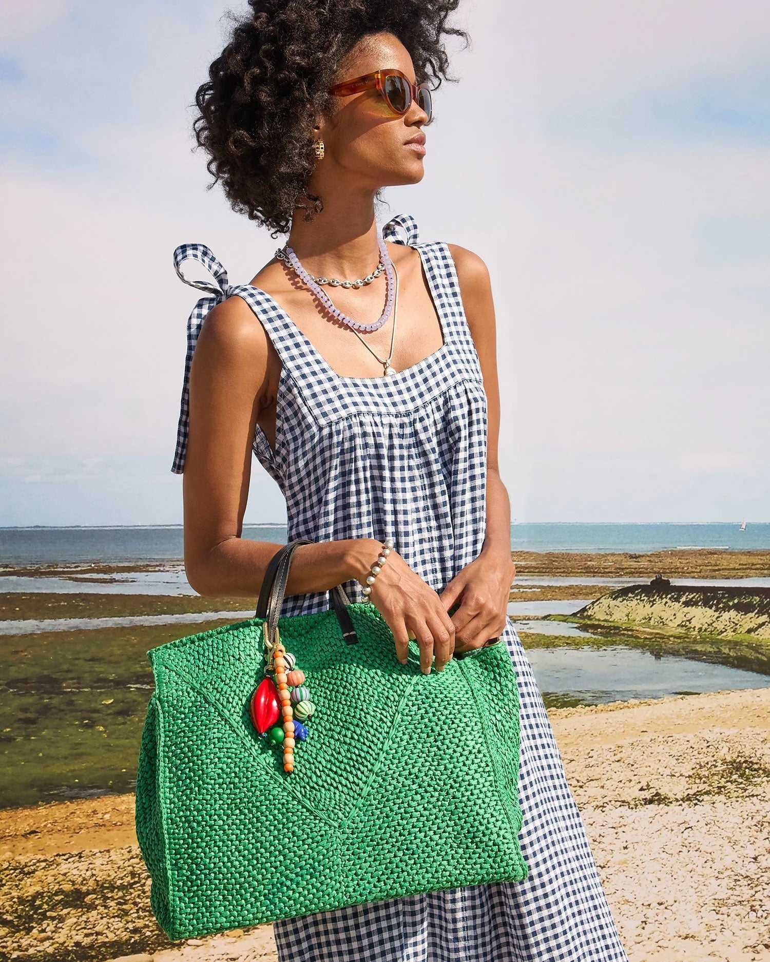 A woman in a black-and-white gingham dress and sunglasses stands on a beach, holding a large green woven bag adorned with the Clare Vivier Stone Slice Necklace. The ocean and sky are visible in the background.