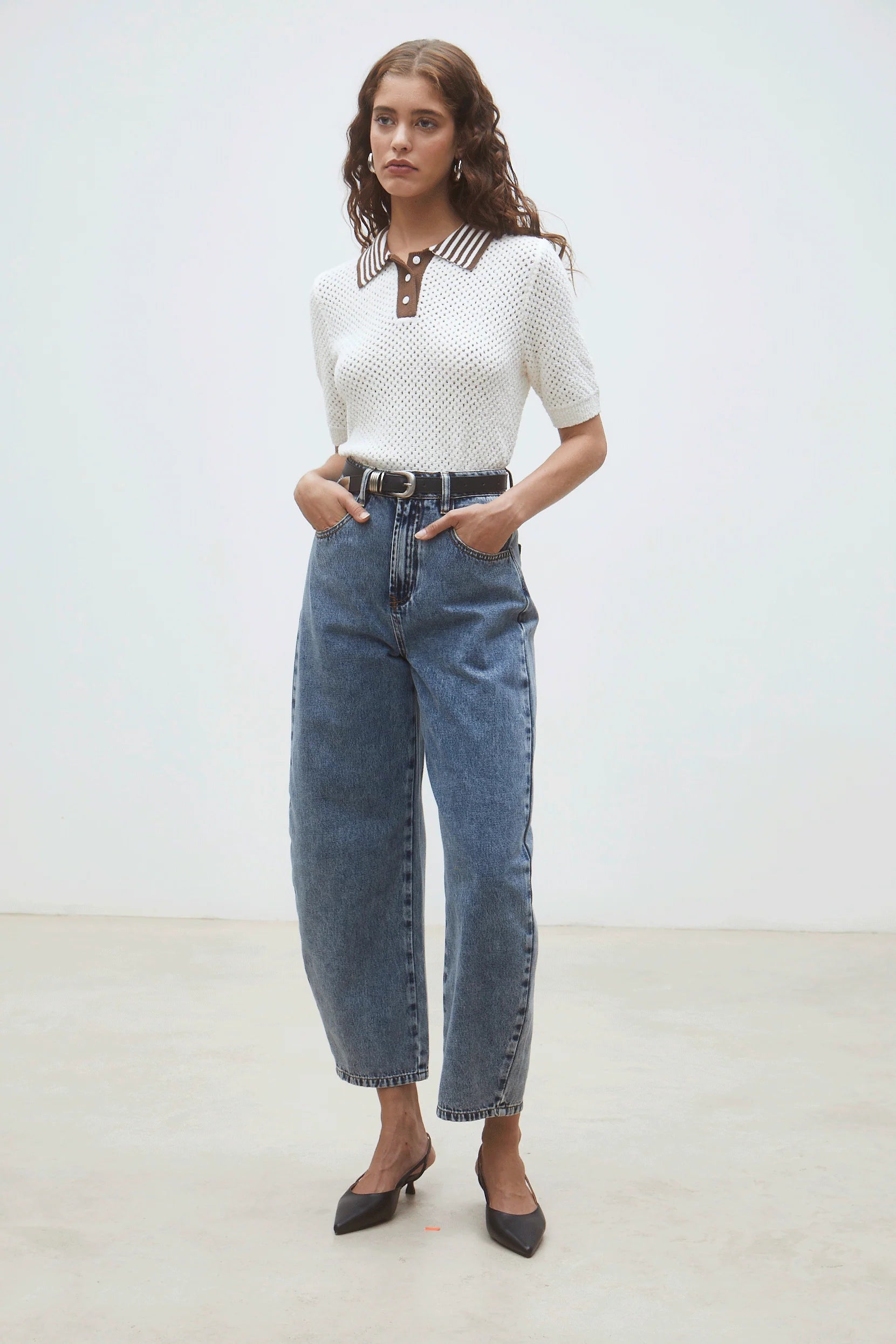 A woman stands against a plain background wearing the SUNCOO Perigue Sweater with a brown and white collar, paired with high-waisted blue jeans, a black belt, and black pointed-toe shoes. Her hands are in her pockets.