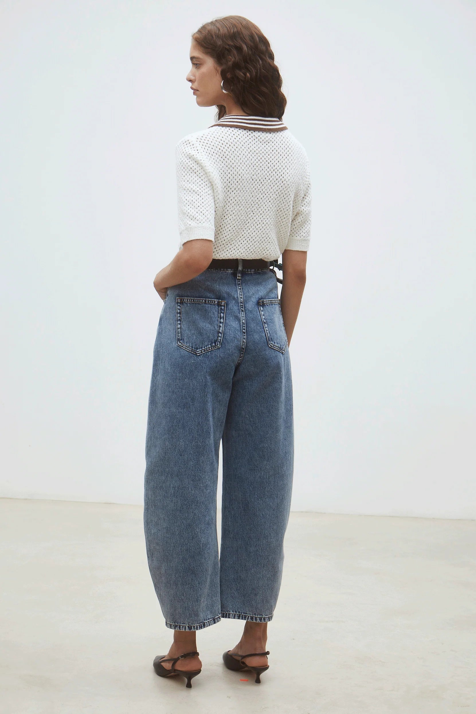 A woman stands facing away indoors against a plain light background, wearing the SUNCOO Perigue Sweater— a white polo-style sweater with a brown and white collar— paired with high-waisted loose blue jeans and black heeled shoes.