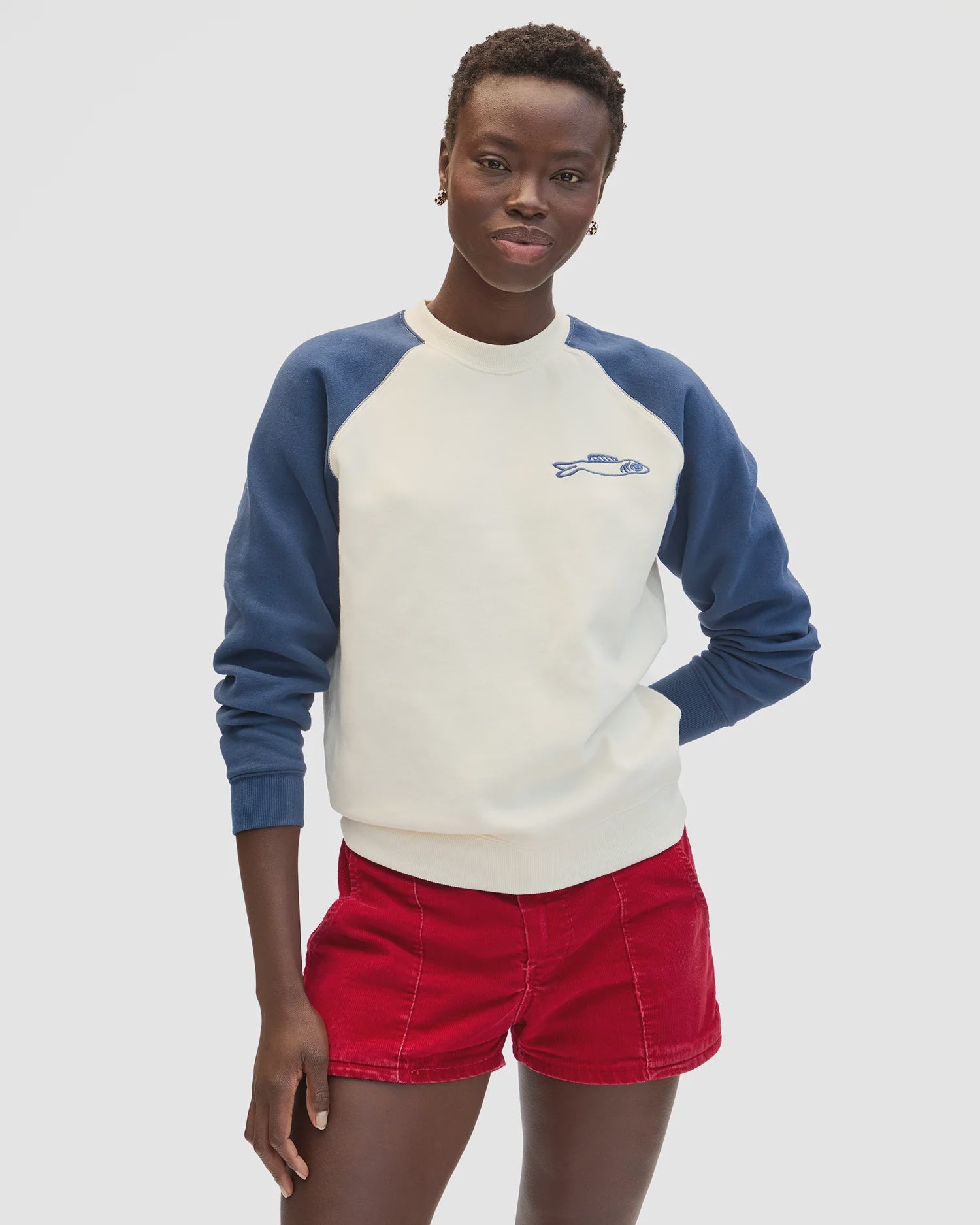 A person wears the Clare Vivier Sweatshirt F25, a cozy cotton fleece with blue sleeves and a small fish graphic, paired with red shorts, standing against a plain white background.