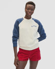 A person wears the Clare Vivier Sweatshirt F25, a cozy cotton fleece with blue sleeves and a small fish graphic, paired with red shorts, standing against a plain white background.