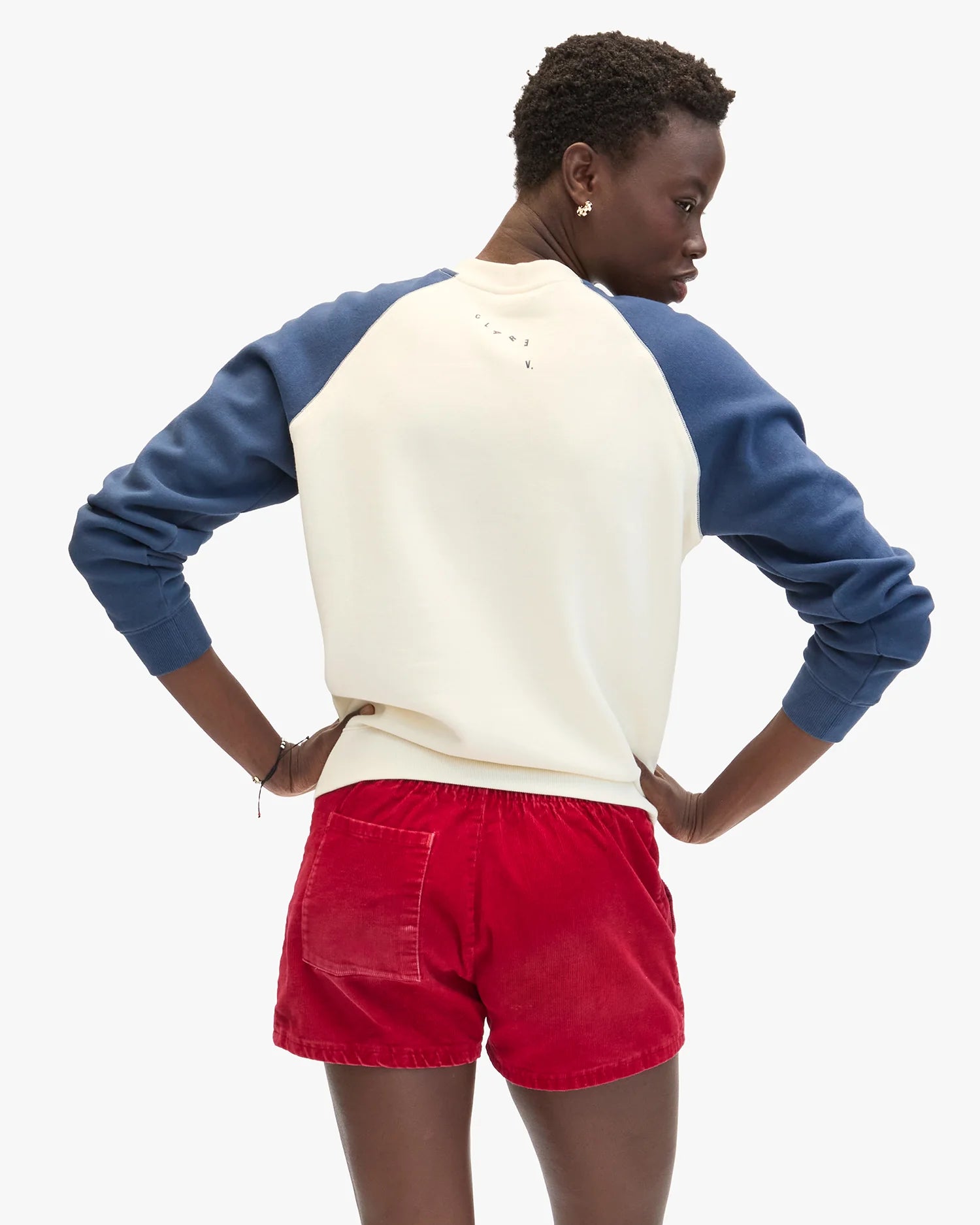 A person stands with hands on hips, wearing the Clare Vivier Sweatshirt F25—cozy cotton fleece with blue sleeves—and red shorts, seen from behind against a plain white background.