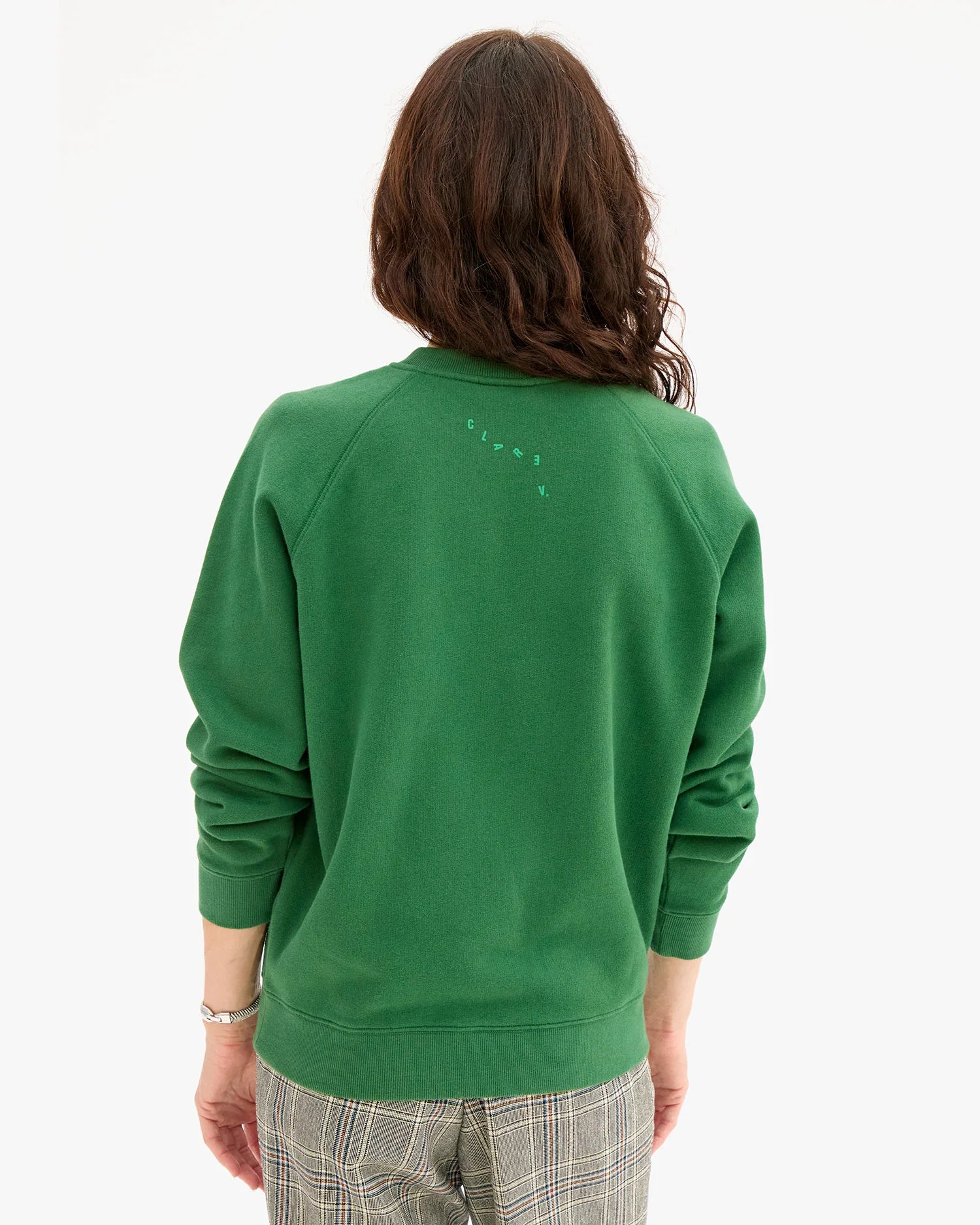A person with wavy brown hair is shown from the back, wearing a green Clare Vivier Sweatshirt / Flocked OUI with small text near the neck and plaid pants. The background is plain white.