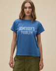 A woman with long, light brown hair wears the DayDreamer Morgan Wallen Somebodies Problem blue tee and olive green pants, standing against a plain beige background.