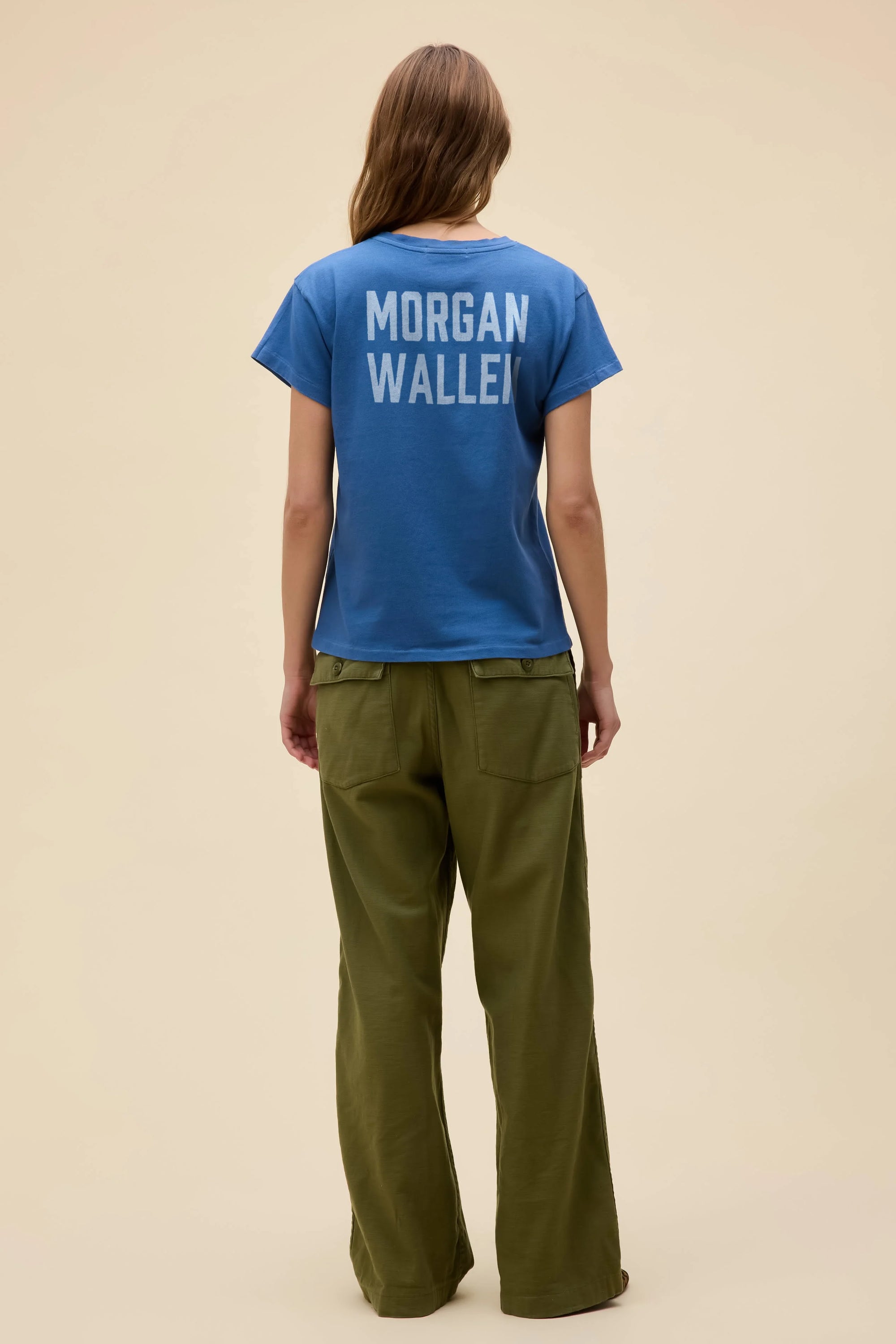 A person with long hair faces away, wearing the DayDreamer Morgan Wallen Somebodies Problem tee in blue and olive green pants against a light beige background, echoing the country star’s iconic style.
