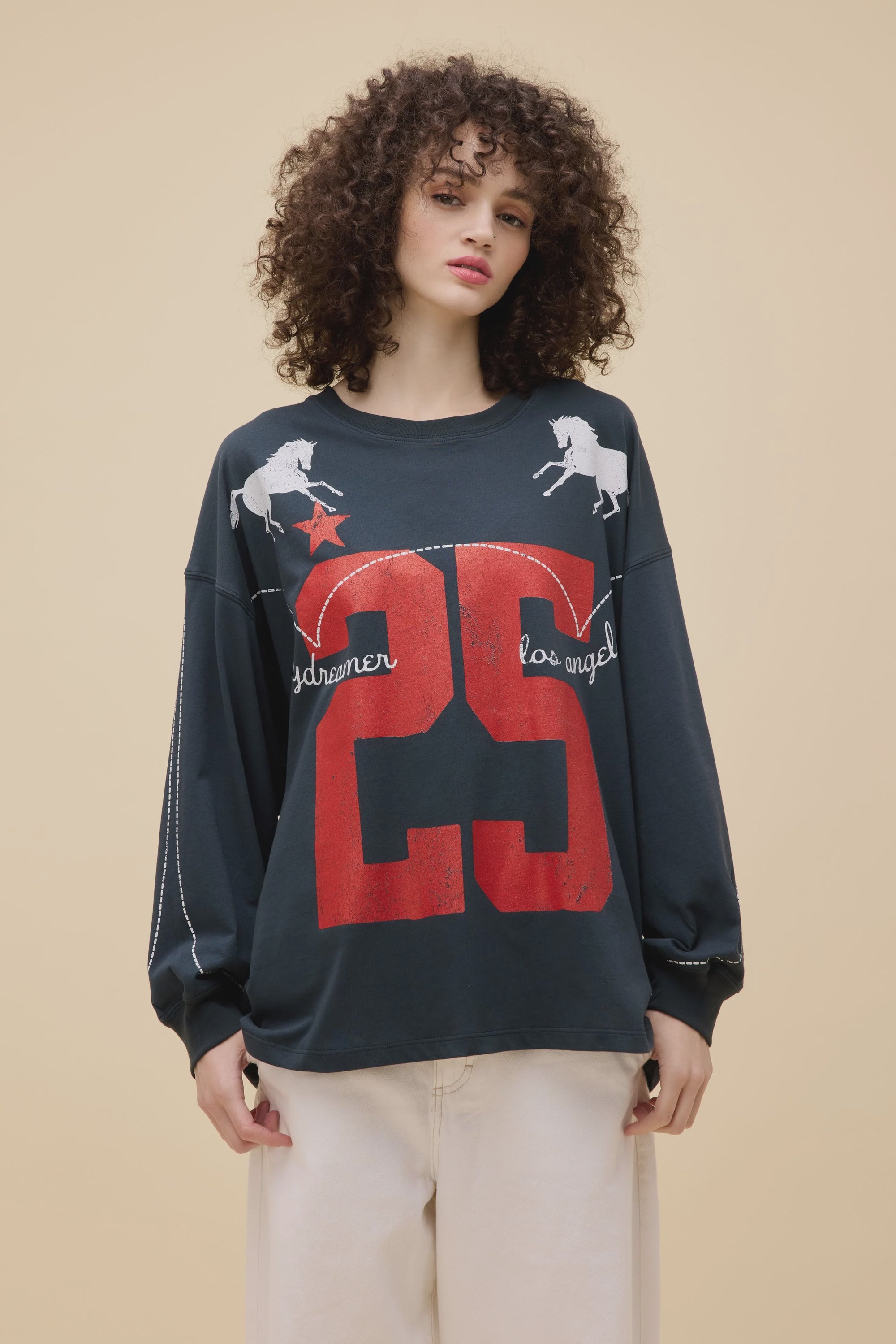 A woman with curly hair wears the DayDreamer DD Los Angeles 25 OS oversized black sweatshirt with western motifs—two white unicorns, a red star, "dreamer," "los angeles," and the number 25—paired with off-white pants against a beige background.