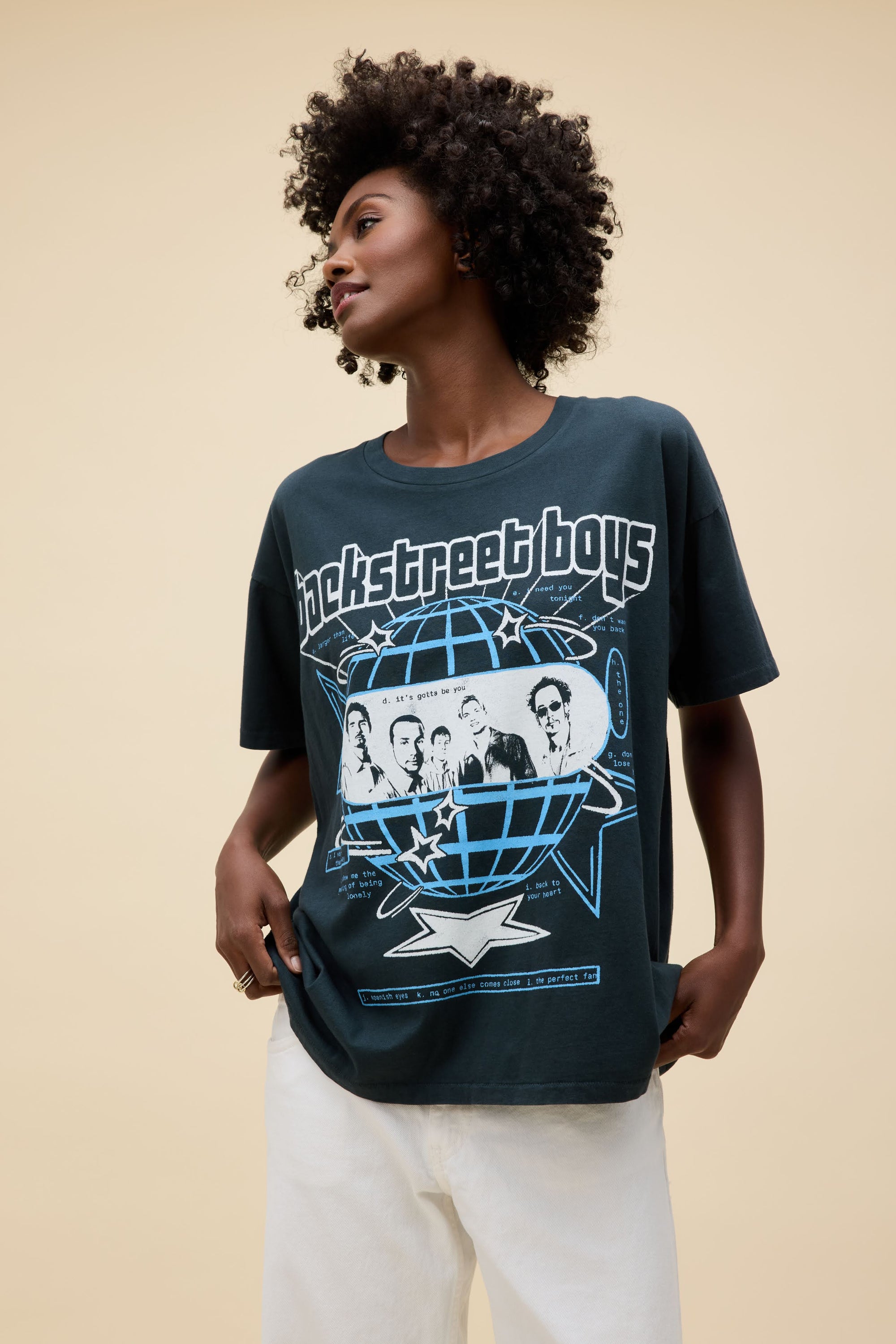A woman with curly hair and white pants wears an oversized DayDreamer Backstreet Boys Into The Millennium tee, featuring band graphics, a globe, and blue stars, standing against a plain beige background.