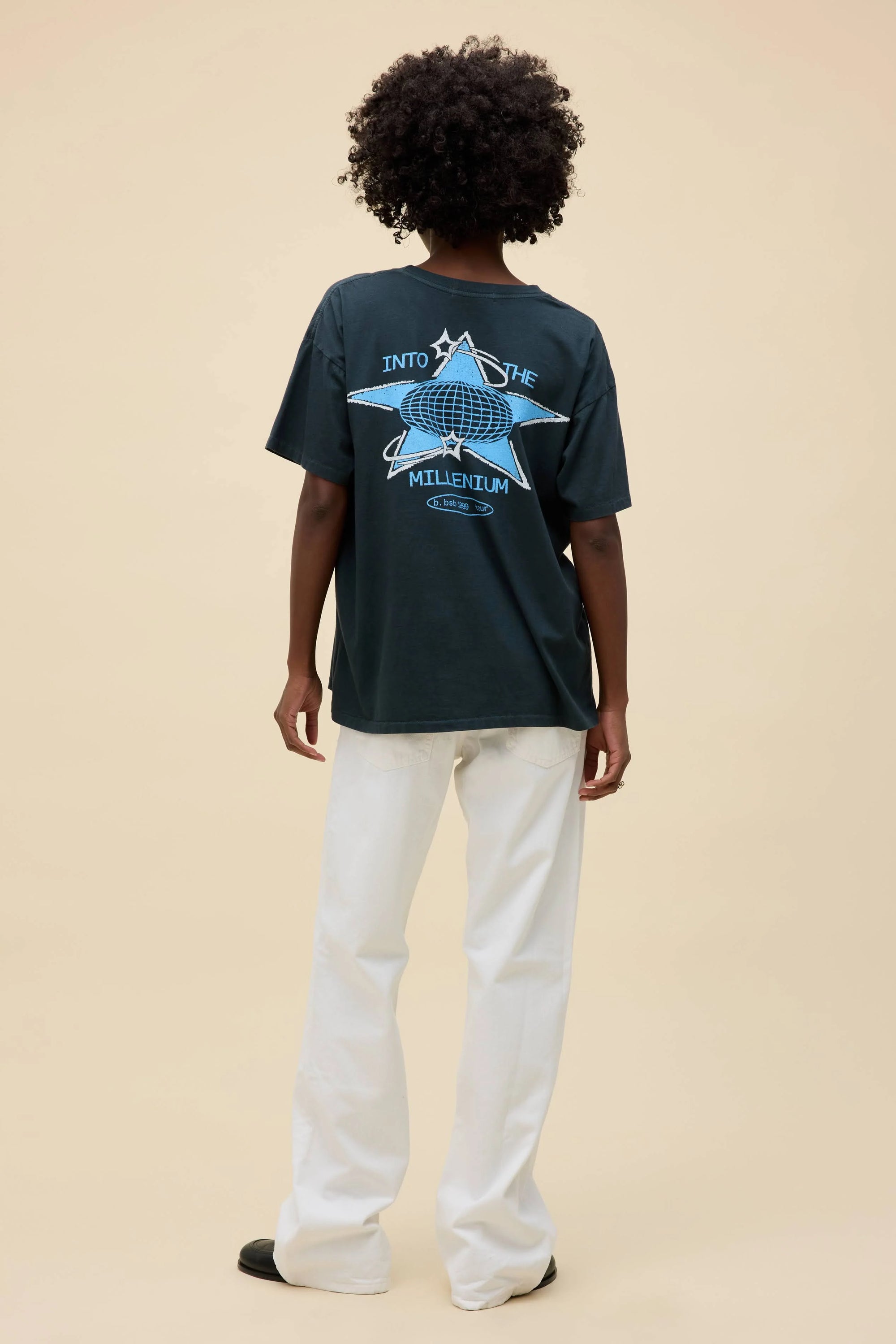 A person with short curly hair faces away, wearing the DayDreamer Backstreet Boys Into The Millennium oversized tee featuring "Into the Millennium" with a globe and star on the back, light pants, and black shoes—a tribute to classic band merch.