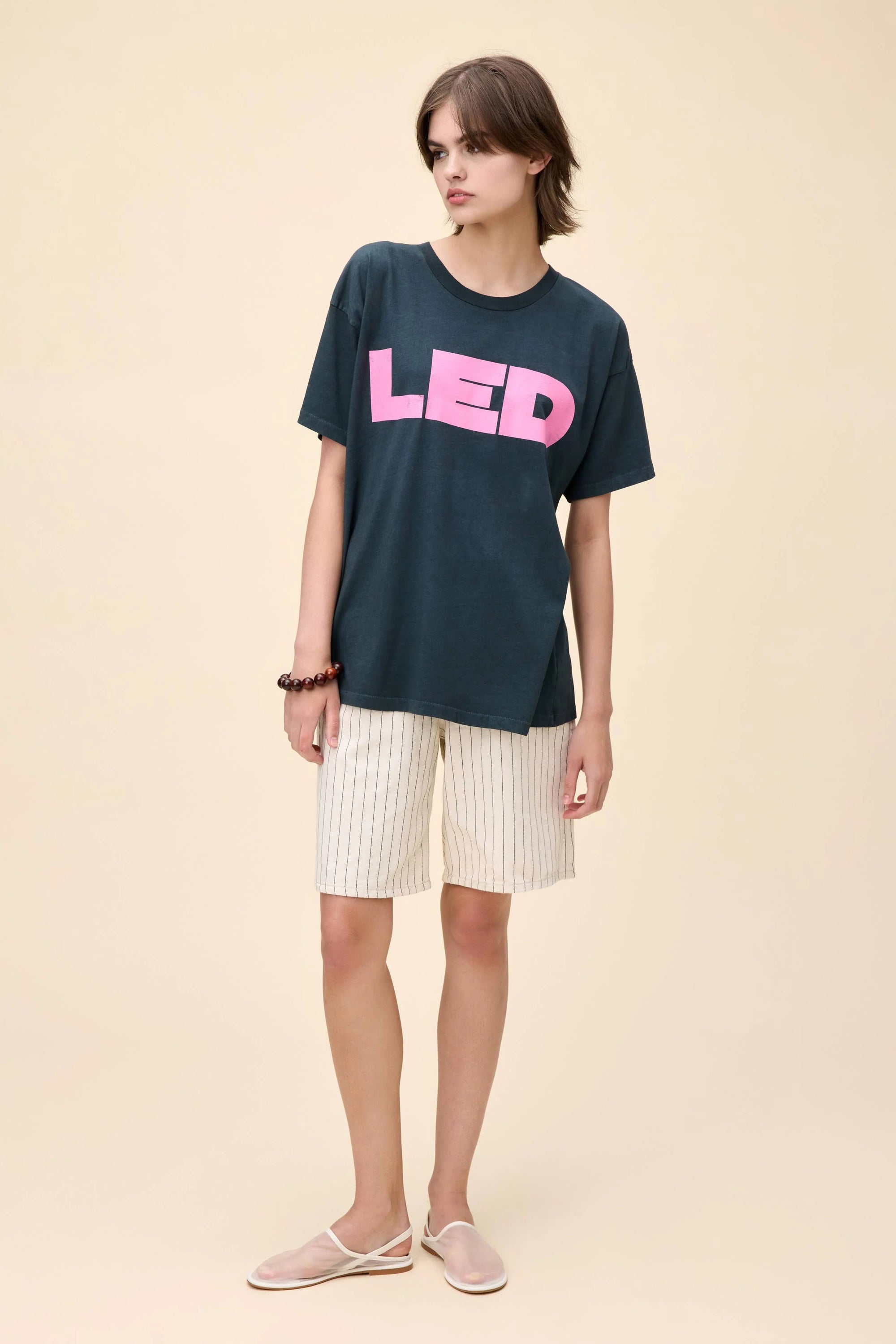 A person wears a dark, oversized Led Zep Merch Tee by DayDreamer with bold pink "LED" lettering, paired with cream striped shorts and sandals, standing against a plain beige background.