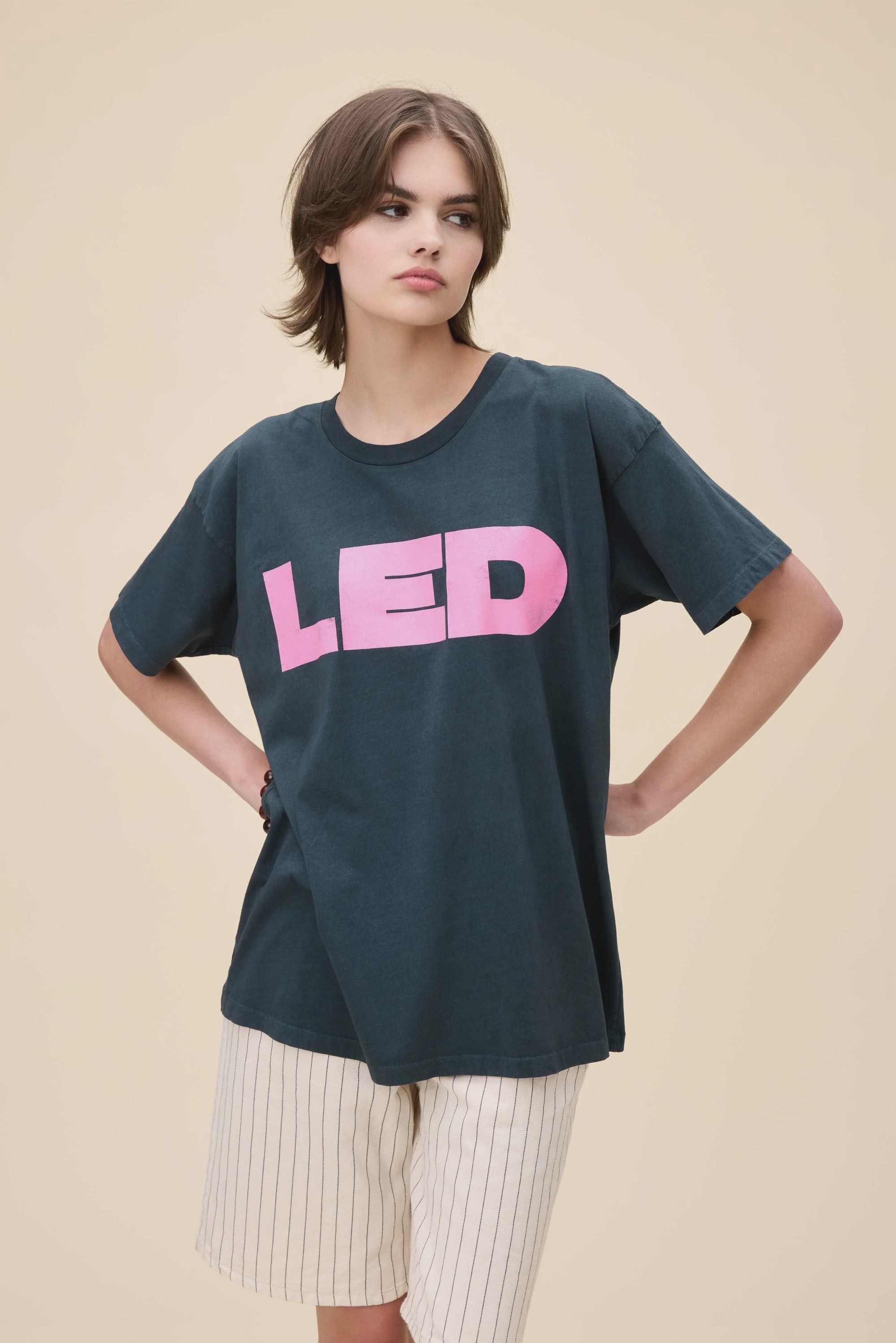 A person with short brown hair wears a DayDreamer Led Zep Merch Tee featuring large pink "LED" letters and cream-striped shorts, standing hands on hips against a plain beige background.