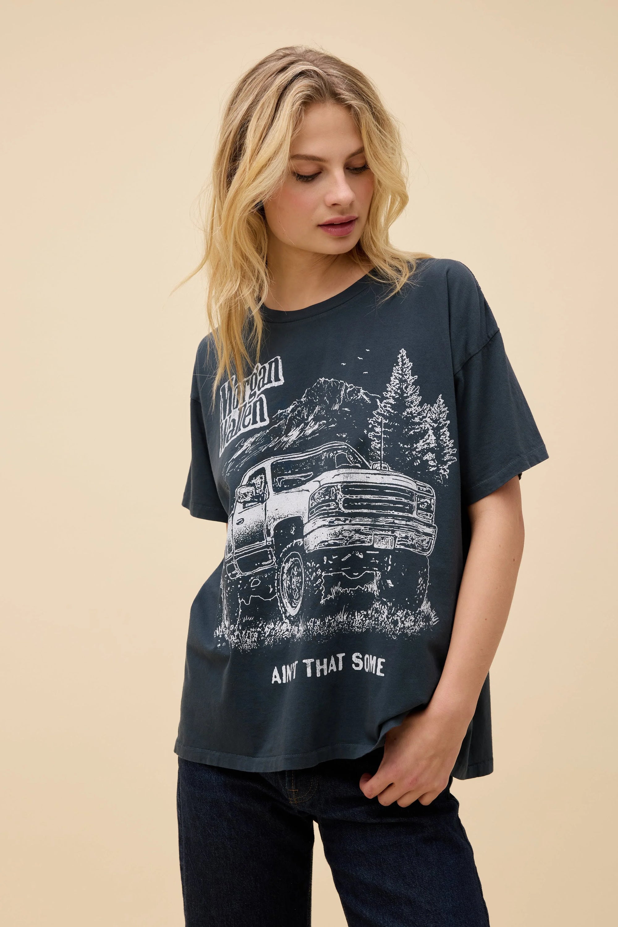 A blonde woman wears the DayDreamer "Morgan Wallen Ain't That Some" oversized cotton t-shirt, which features a pickup truck, trees, mountains, and the text "Morgan Wallen Ain't That Some." She gazes downward with one hand at her side.