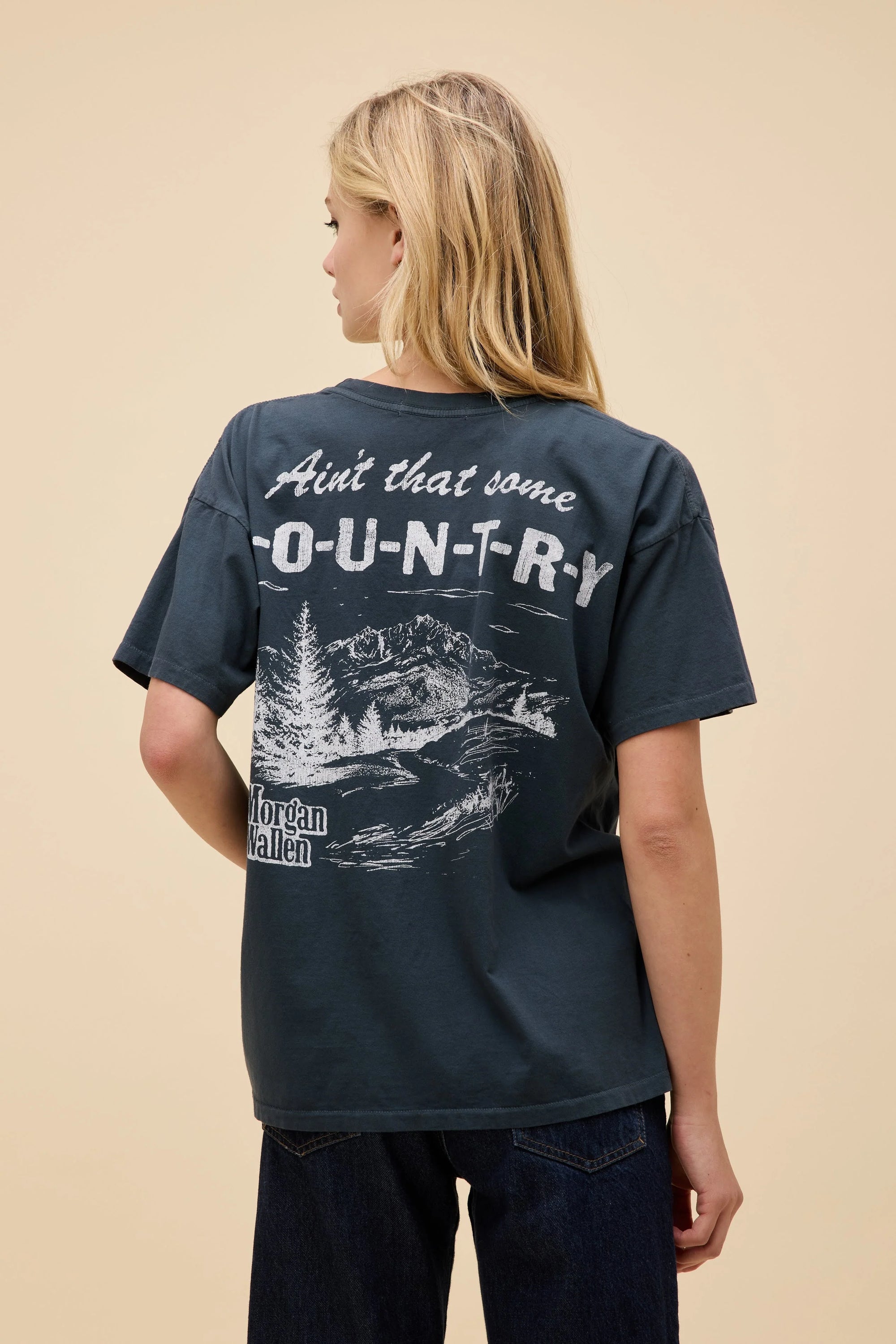 A woman with long blonde hair wears the DayDreamer Morgan Wallen Ain't That Some oversized cotton t-shirt, featuring white text and a scenic back graphic—perfect Morgan Wallen merch for fans. She faces away from the camera.