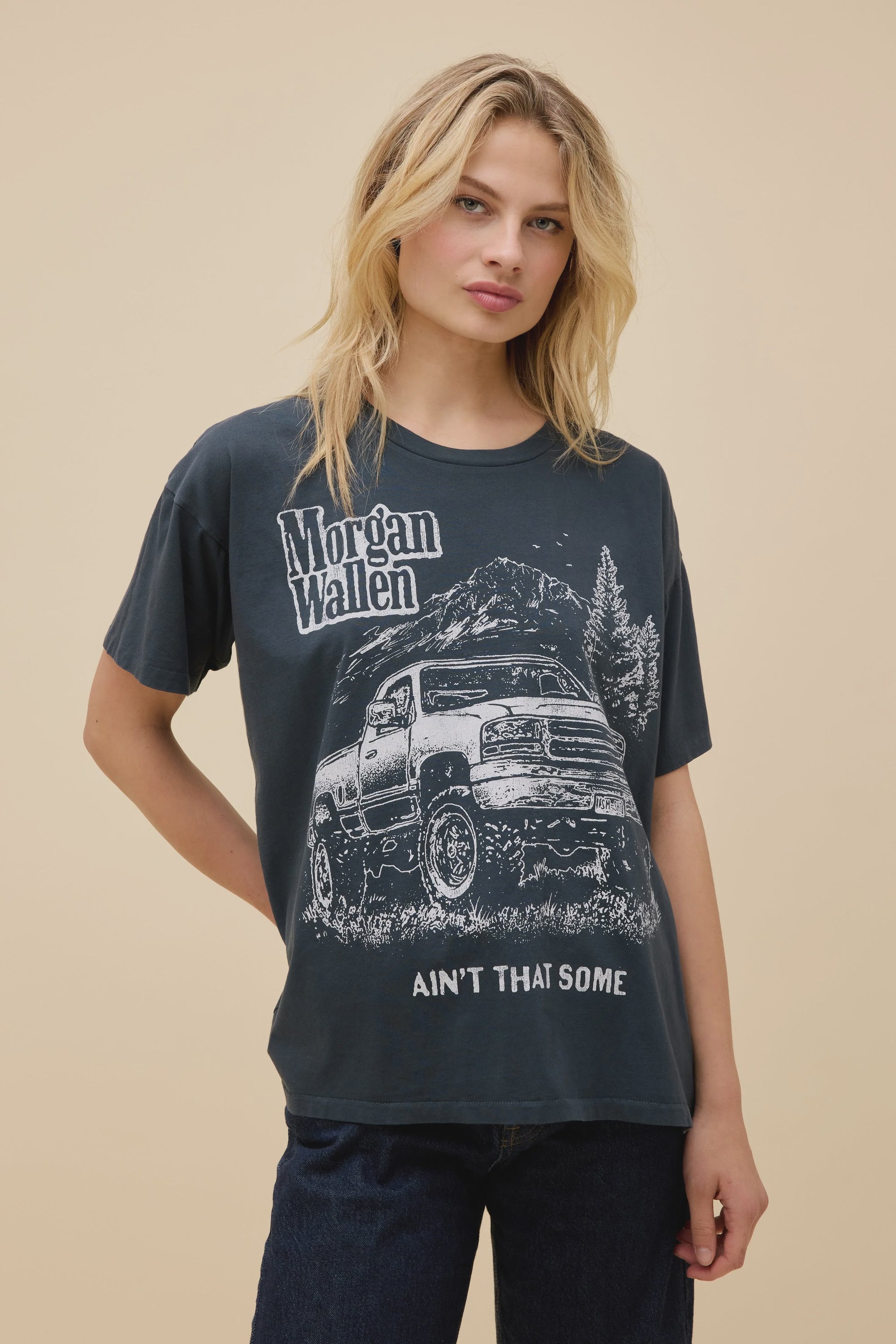 A blonde woman models the DayDreamer Morgan Wallen Ain't That Some oversized cotton T-shirt, which features a truck, trees, and the phrase "Ain't That Some." She stands against a plain beige background, looking slightly to the side.