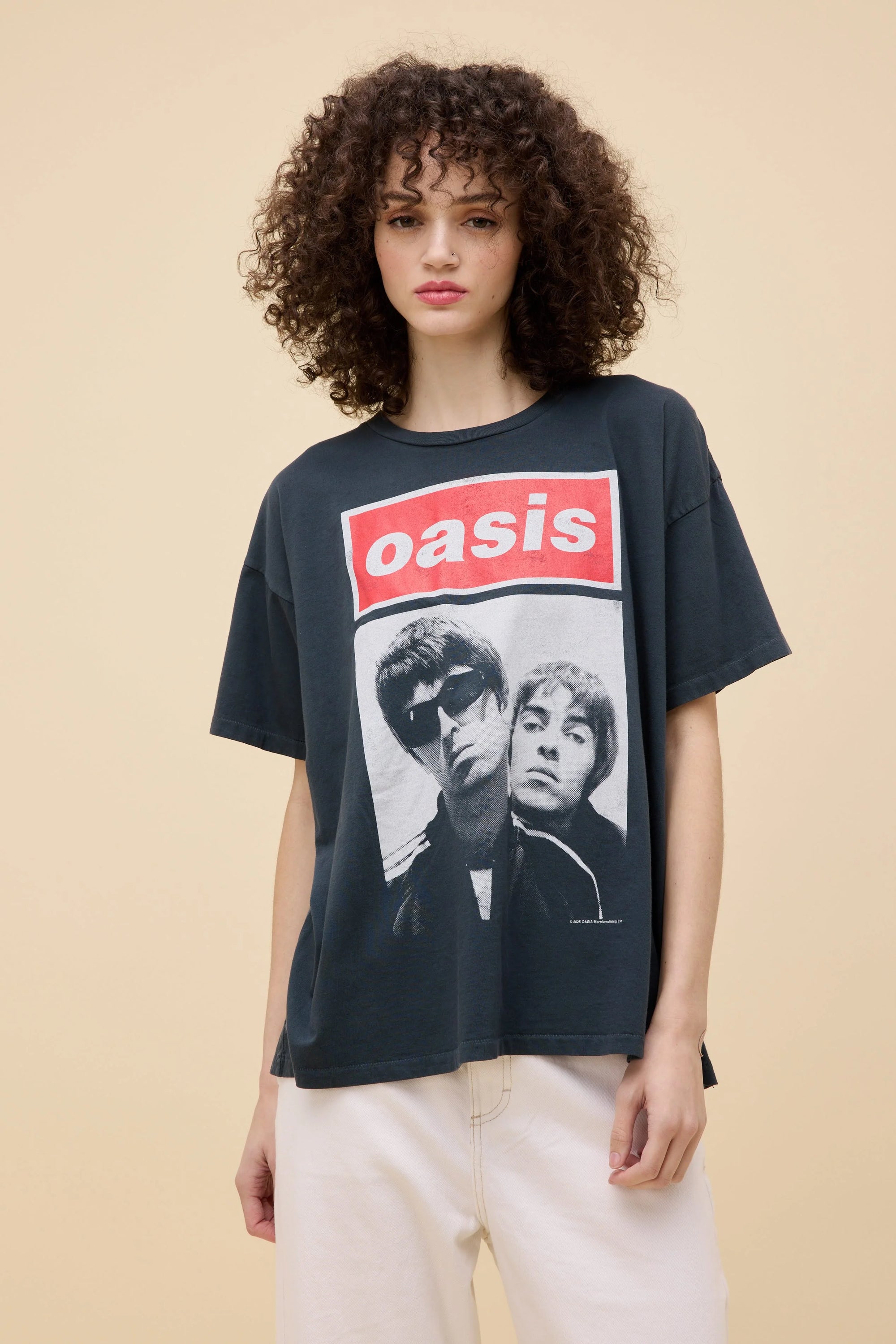 A young woman with curly hair wears the DayDreamer Oasis Merch Tee—an oversized black shirt with a photo of two men and the band's name in bold red letters—paired with light-colored pants, against a beige background.