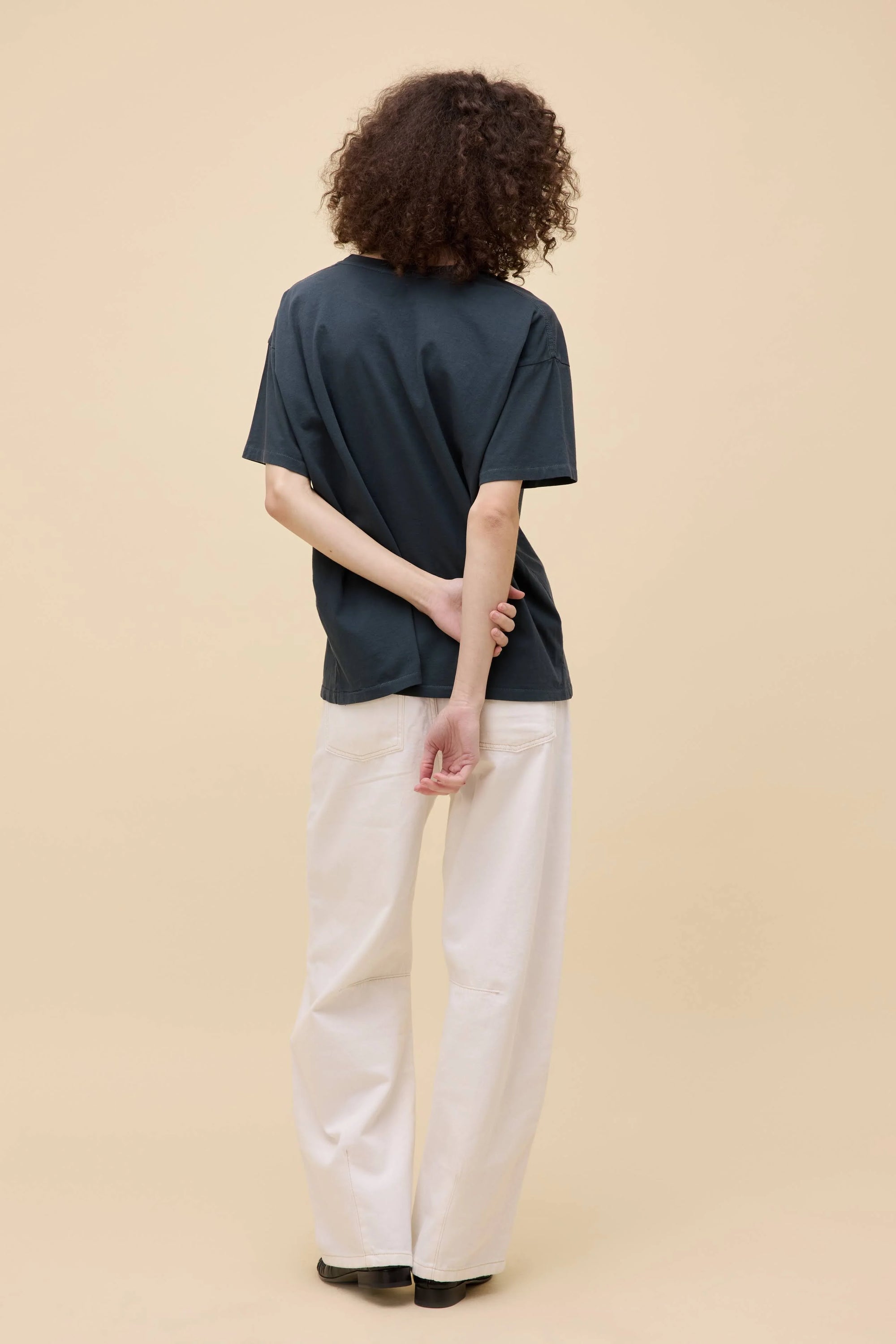 A person with curly hair faces away, wearing the oversized Oasis Merch Tee by DayDreamer, paired with white pants and black shoes, one hand behind their back against a neutral beige background.
