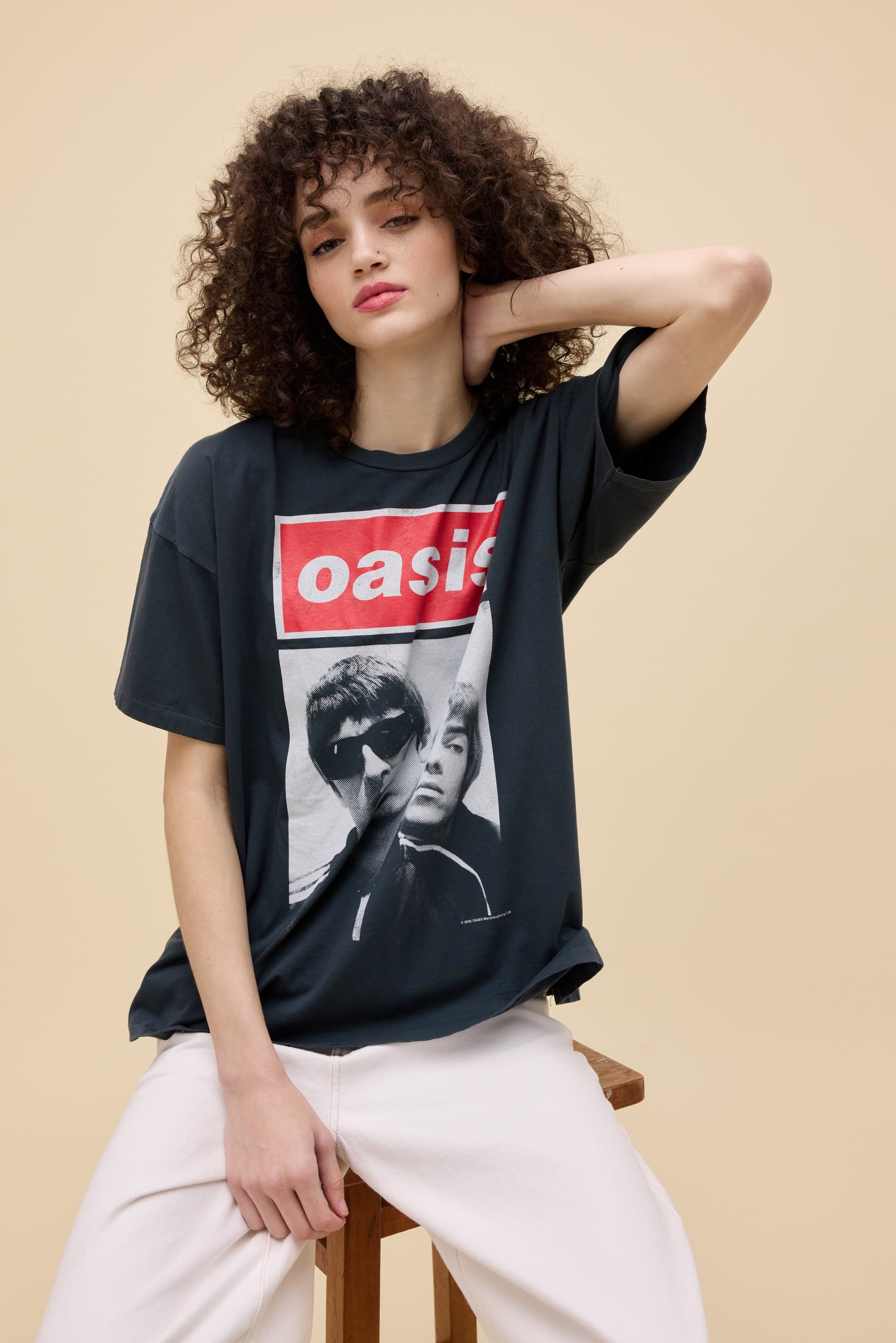 A person with curly hair wears the oversized DayDreamer Oasis Merch Tee and light pants while sitting on a wooden stool against a beige background, resting one hand behind their head.