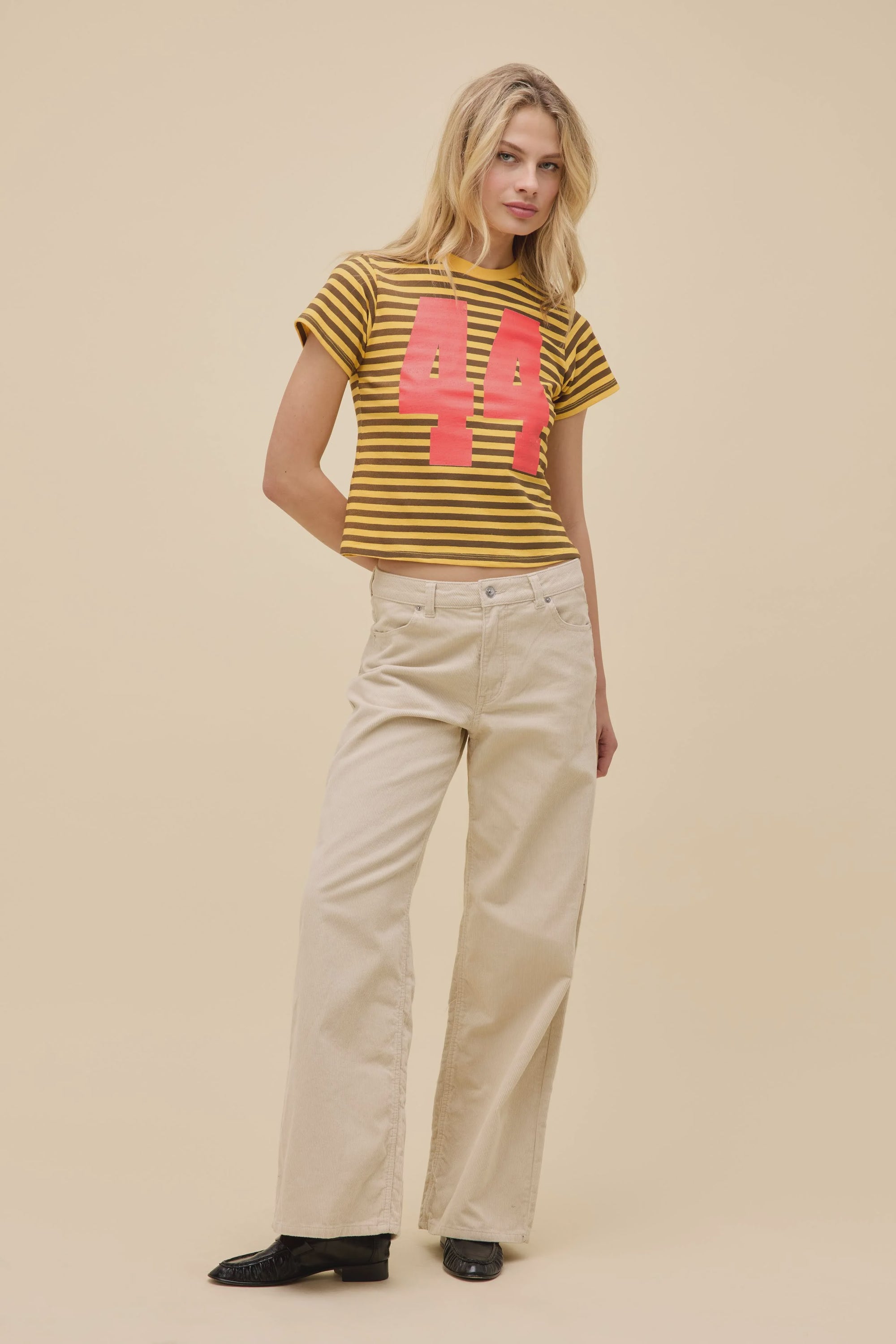 A woman poses against a beige background wearing DayDreamer's 44 Striped Vintage Tee, featuring a bold pink "44," styled with cream wide-leg pants and black shoes for a retro-inspired look.