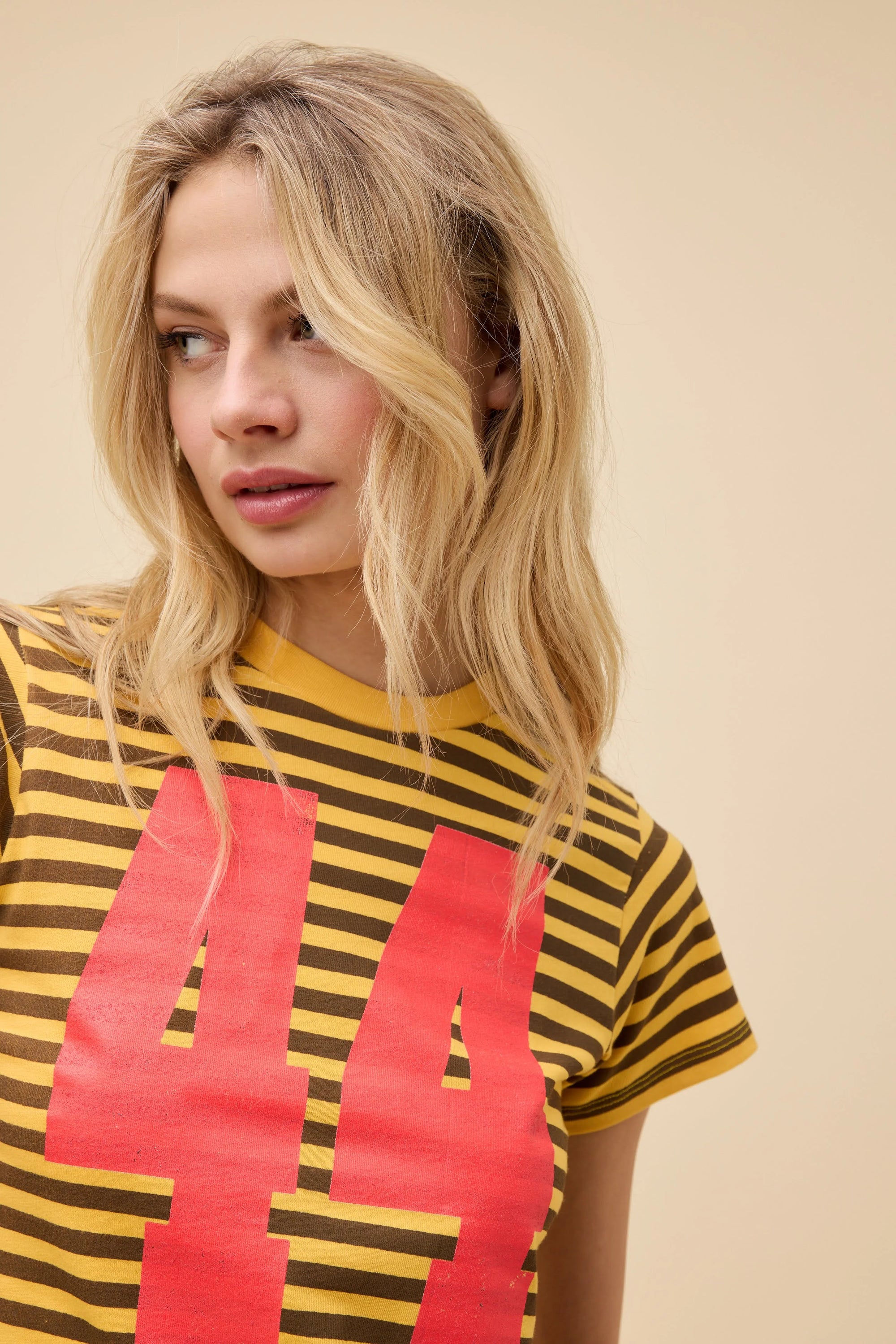 A woman with wavy blonde hair wears the DayDreamer 44 Striped Vintage Tee in yellow and brown with a bold red "44" on the front, standing against a plain beige background and looking to her left.