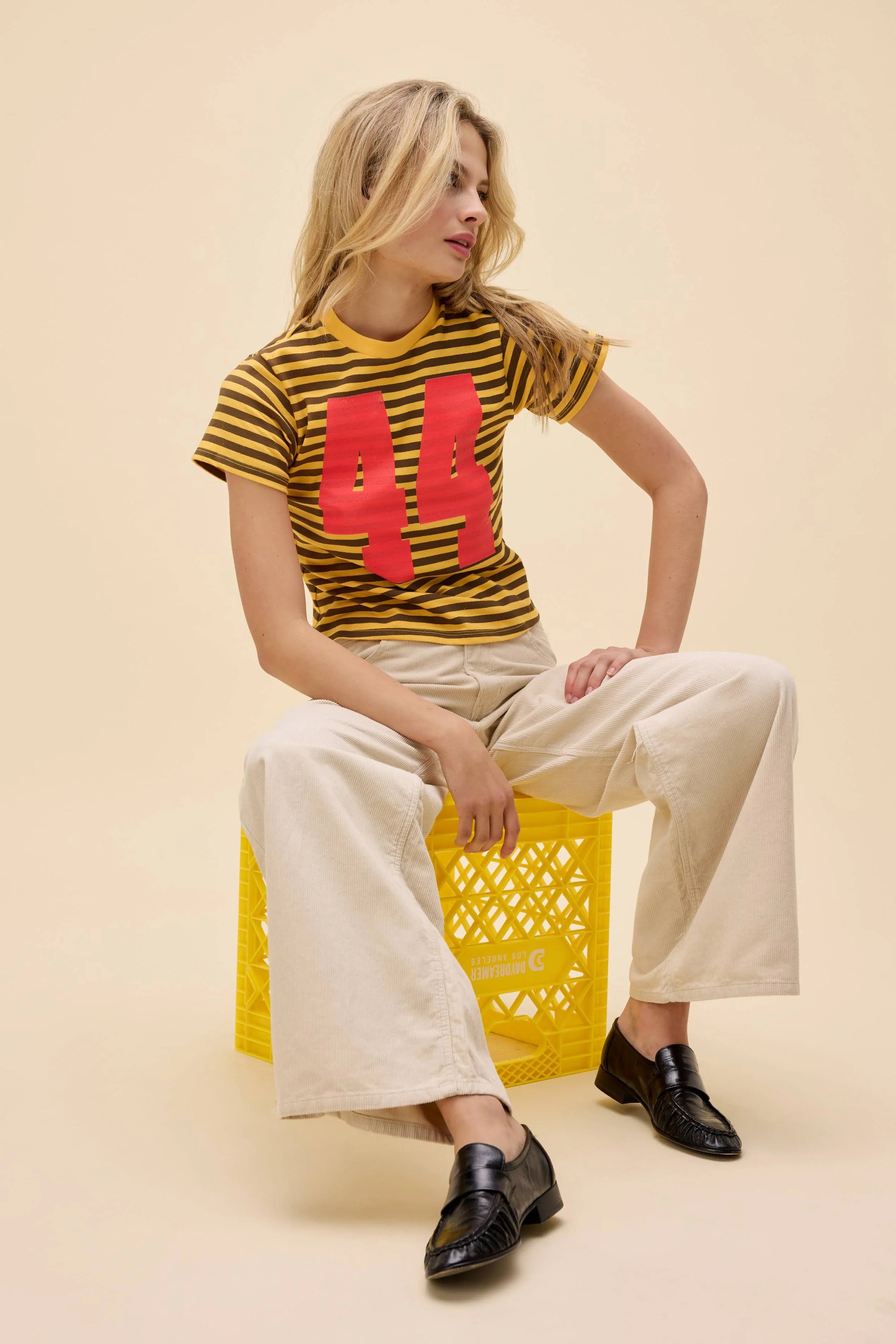 A woman with long blonde hair sits on a yellow crate, wearing the DayDreamer 44 Striped Vintage Tee, cream wide-leg pants, and black loafers. She looks to her left against a beige background.