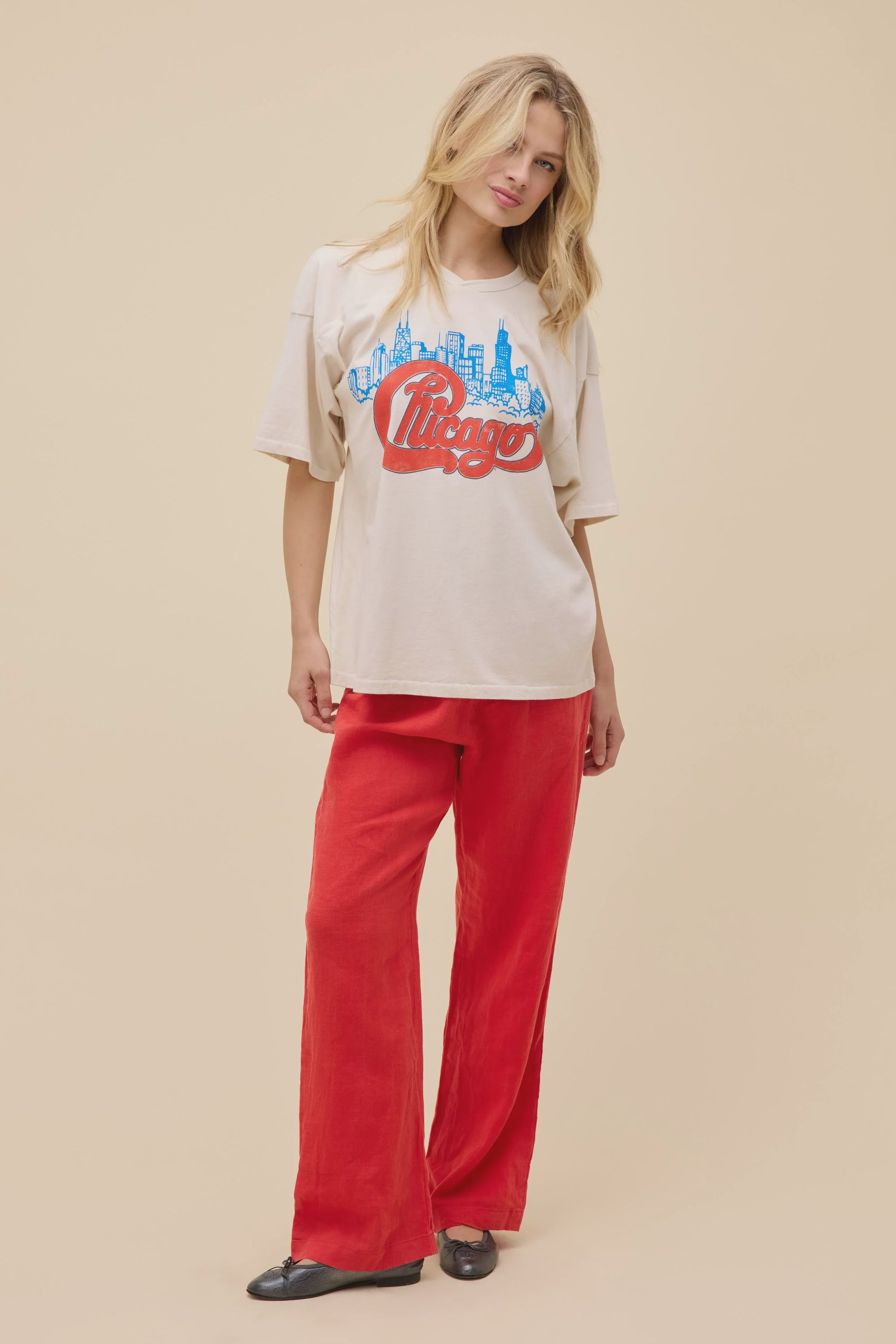 A person wears the DayDreamer Chicago Skyline Tee, featuring a blue skyline graphic and red "Chicago" text, paired with loose red pants and black shoes, standing against a plain beige background.