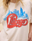 A person wearing the DayDreamer Chicago Skyline Tee sports a white shirt with a blue skyline outline, bold red cursive "Chicago," and is made from heavyweight cotton for lasting comfort.