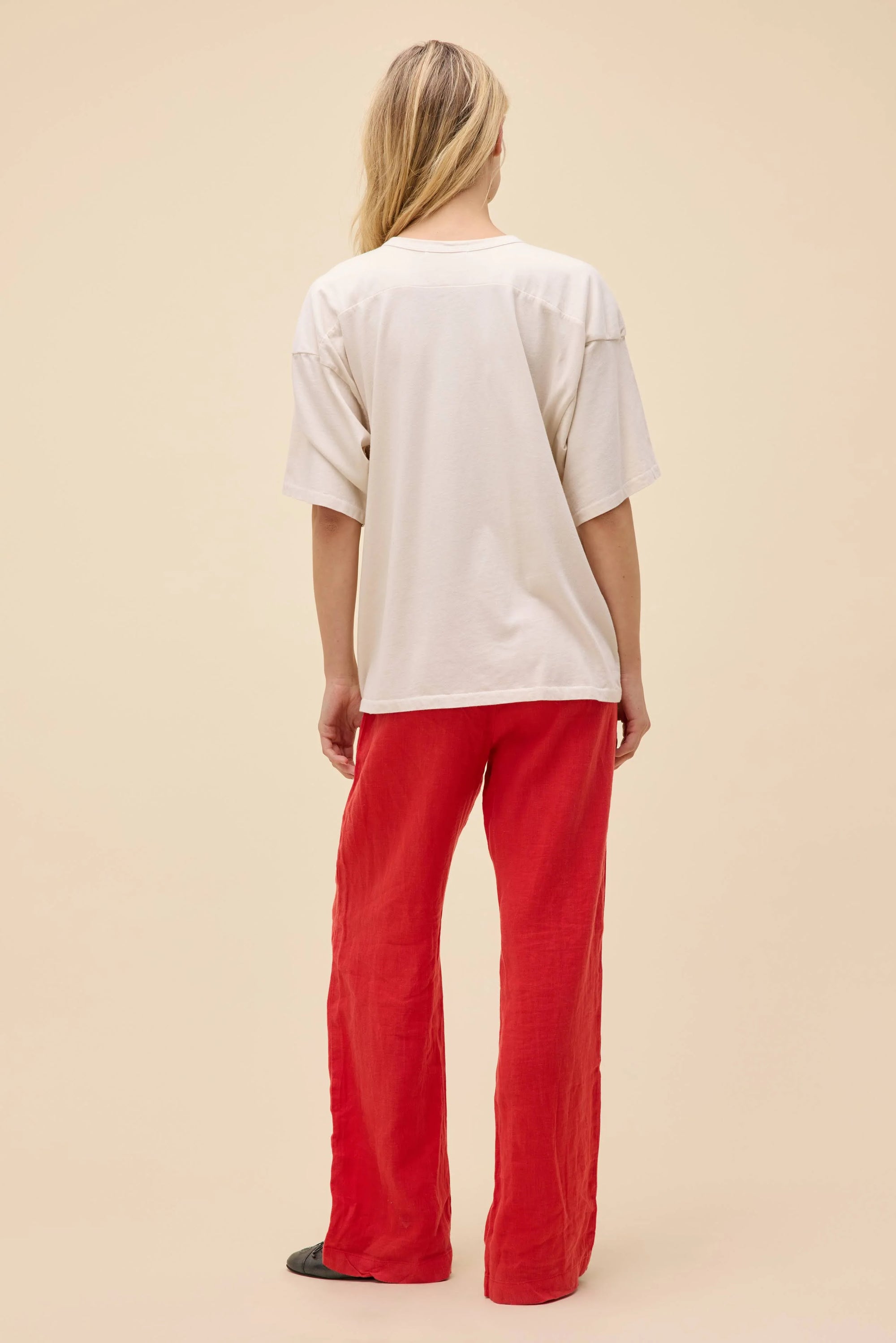 A person with long blonde hair, seen from behind, wears the DayDreamer Chicago Skyline Tee, bright red pants, and black shoes, standing against a plain beige background.