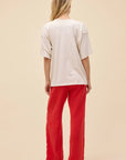 A person with long blonde hair, seen from behind, wears the DayDreamer Chicago Skyline Tee, bright red pants, and black shoes, standing against a plain beige background.