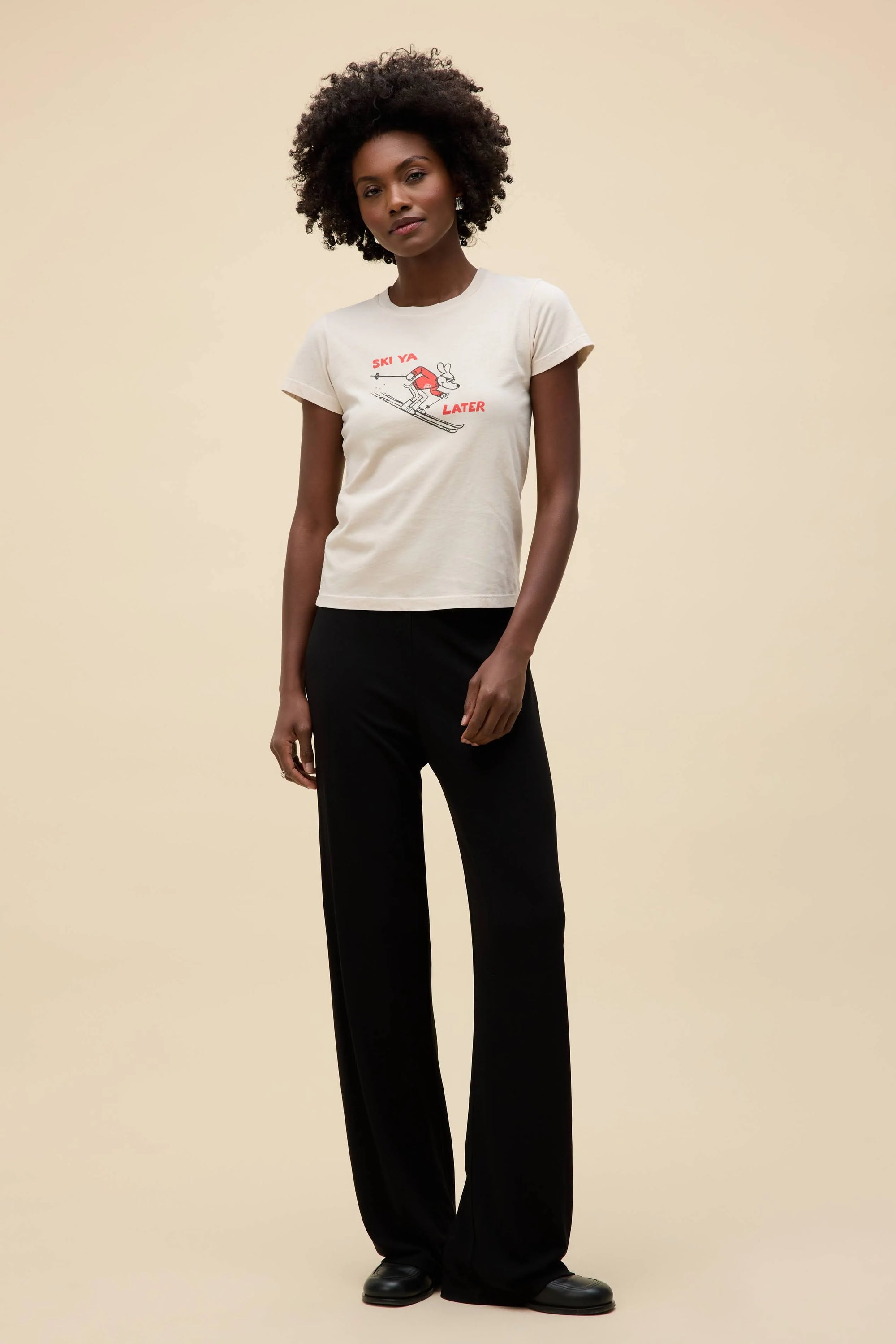 A woman with short curly hair wears DayDreamer's Ski Ya Later Tee and black wide-leg pants, standing against a plain beige background.