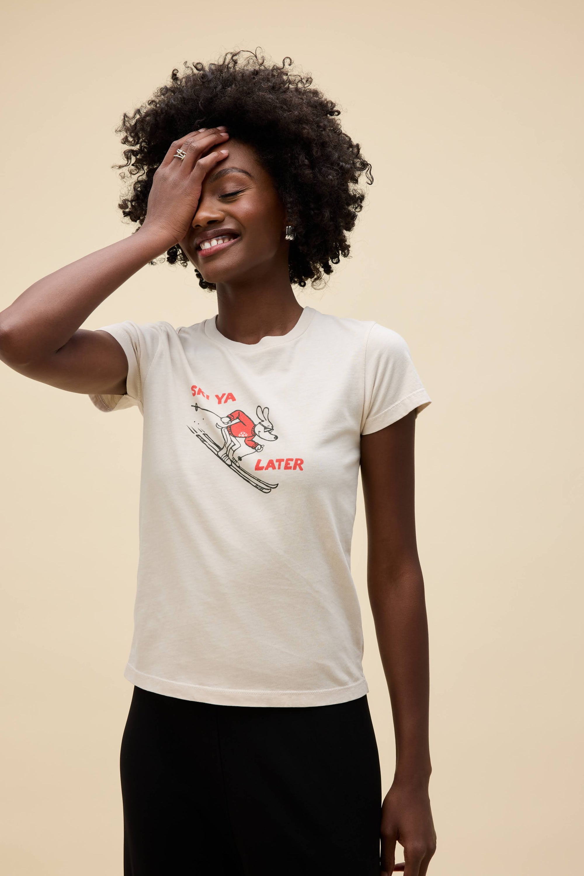 A smiling woman with curly hair stands against a beige background in the DayDreamer Ski Ya Later Tee—a women’s cotton t-shirt featuring a playful graphic and the words “SKI YA LATER.” She covers part of her face with one hand, looking relaxed and happy.