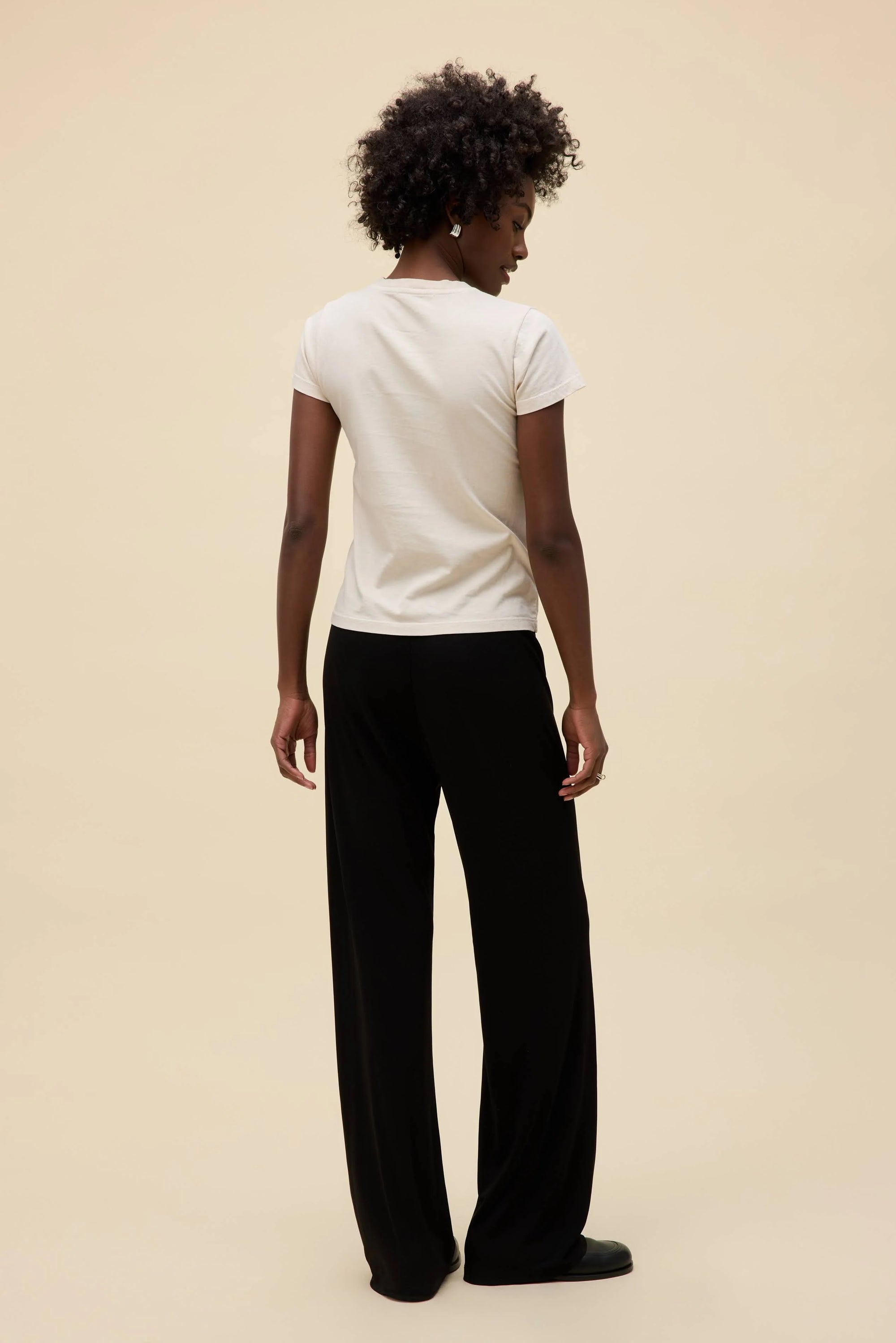 A person with short curly hair stands facing away, wearing the DayDreamer Ski Ya Later Tee, loose black pants, and black shoes against a plain beige background.