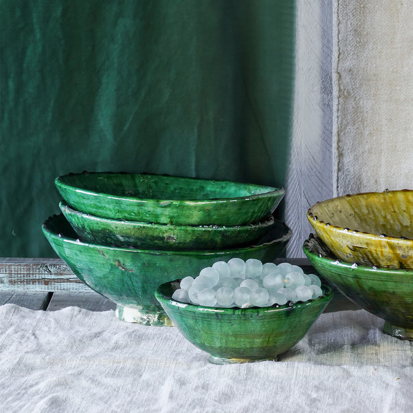 A set of hand-shaped ceramic bowls from Blue Ocean Traders, featuring mostly green and one yellow Tamegroute Bowl, are stacked on a white cloth. The smallest bowl in front displays round, translucent white beads as decoration.