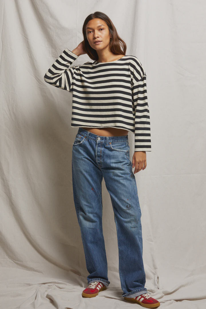 A person with long walnut-colored hair wears a Taylor Long Sleeve Tee by Perfectwhitetee in black and white stripes, paired with blue jeans and red sneakers. They stand against a light fabric backdrop, casually resting one hand on their head.