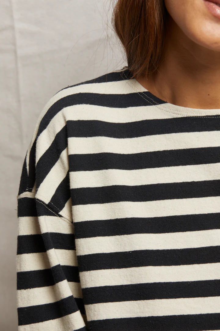 A person wears the Taylor Long Sleeve Tee by Perfectwhitetee, a black-and-white striped shirt crafted from recycled cotton, shown from neck to chest. It has wide horizontal stripes with long hair partially visible on the right against a walnut-shaded neutral background.